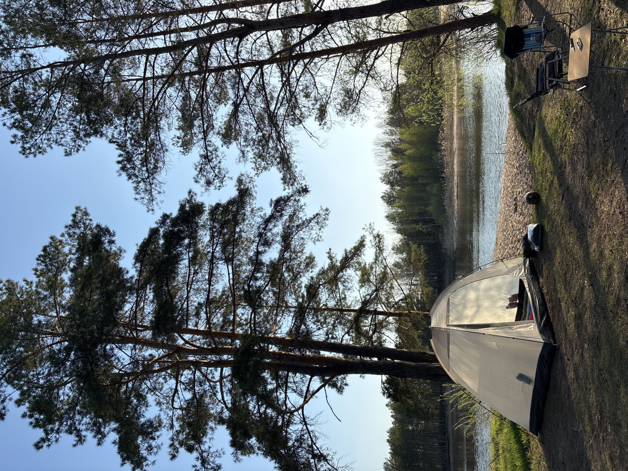 The image shows a campsite on the bank of a body of water, likely a lake or river, in what appears to be a wooded area. A tent is pitched near the water's edge, and trees frame the view. Camping chairs and a small table are set up nearby, suggesting a relaxation or activity area. The location is Vekoshinka, Russia. The scene conveys a sense of leisure and outdoor recreation.