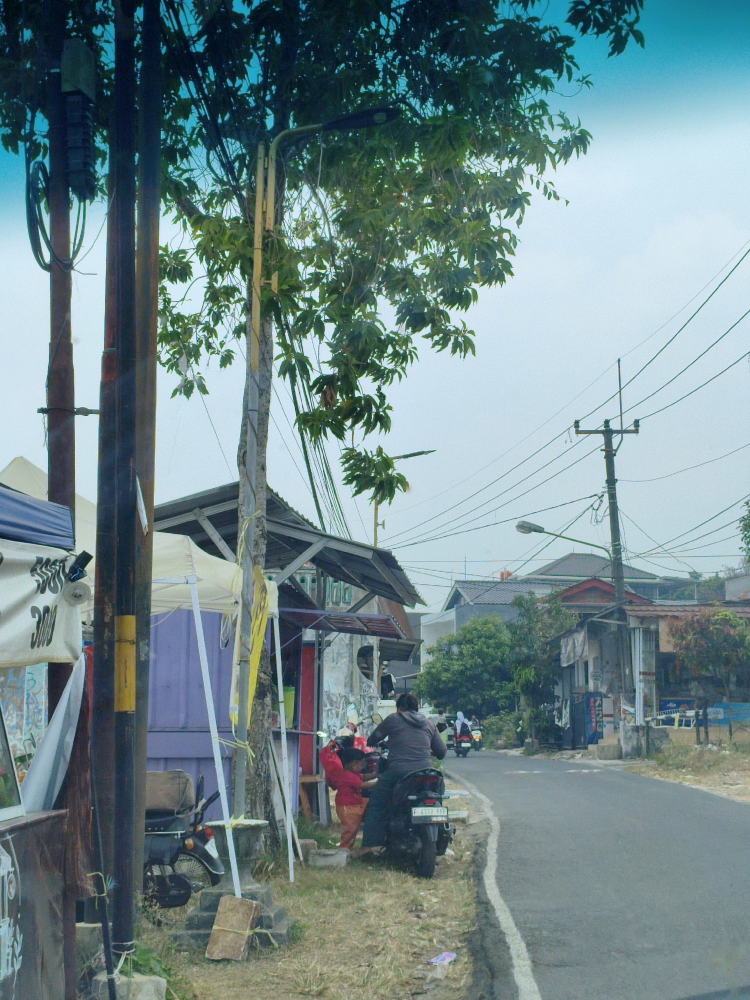 The image depicts a street scene in Sukaraja, Indonesia. In the foreground, a person is sitting on a motorcycle parked on the grassy shoulder of a paved road. The motorcycle has a license plate with the text "F 2932 FEB". Behind the motorcycle, a child wearing a red top and orange pants is standing, facing the person on the motorcycle, possibly interacting with them. A tree with green leaves is partially visible in the upper left of the frame, with its branches extending over a wooden pole and electrical wires. Several tents and awnings are set up along the side of the road, suggesting a temporary market or roadside stalls. In the background, there are buildings and more trees, with power lines crisscrossing the sky. Further down the road, other motorcycles are visible, indicating traffic. The overall environment appears to be a mix of urban and rural elements, common in Indonesian towns.