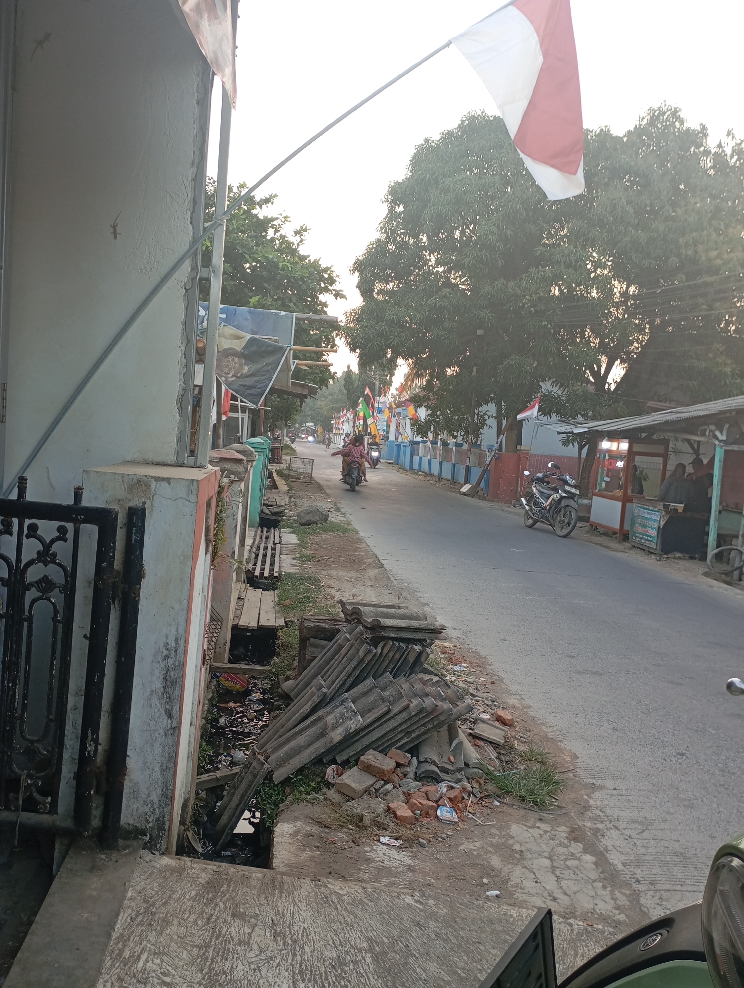 The image depicts a street scene in Kecamatan Haurgeulis, Indonesia, characterized by a slightly overcast sky and warm, late afternoon lighting. In the foreground, to the left, is the exterior wall of a building, partially textured and painted white, with a dark-colored metal gate. Adjacent to the wall is a drainage ditch filled with debris and stagnant water. Piled on the side of the road are stacks of old roofing tiles and broken bricks.
The street itself is paved and appears to be a two-lane road. Several motorcycles are present: one is parked on the right side of the street, near a small food stall or warung with red and white trim and bright interior lighting, where a few individuals are seated. Another motorcycle is in motion on the left side of the road, carrying two people. Further down the street, more motorcycles are visible, some carrying passengers.
Along the sides of the road, there are flags and banners, suggesting a local celebration or national event; the Indonesian flag (red and white) is prominently displayed on a pole. Trees with dense foliage line the street, and there are other buildings and structures visible in the background, including a long, low building with blue and red accents. The overall impression is of a lived-in, somewhat cluttered, and active street in a semi-urban or rural Indonesian setting.