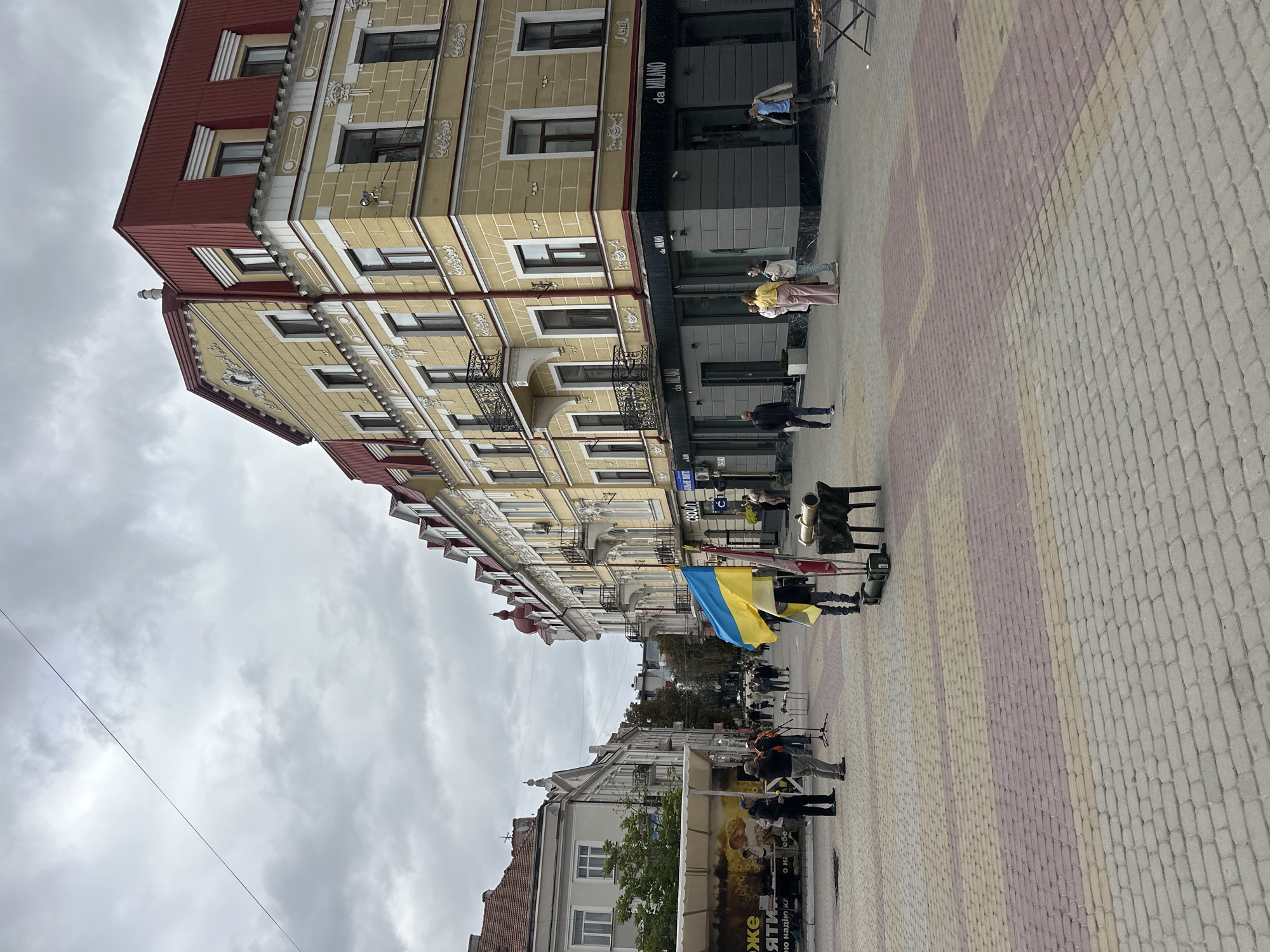 The image depicts a street scene in Ternopil, Ukraine, on an overcast day. The foreground is dominated by a patterned pedestrian walkway paved with interlocking bricks in shades of gray, pink, and yellow. In the mid-ground, a group of people are gathered near a small cannon replica and a stand displaying flags. Two individuals are holding up a Ukrainian flag. To the right of this group, several people are seen walking along the sidewalk in front of a building with dark storefronts labeled "da MILANO". The building itself is a multi-story structure with yellow facades and a red roof. Further down the street, more people are visible, and on the left, another building with a white facade and a white canopy can be seen. The sky is heavily clouded, suggesting potential rain.