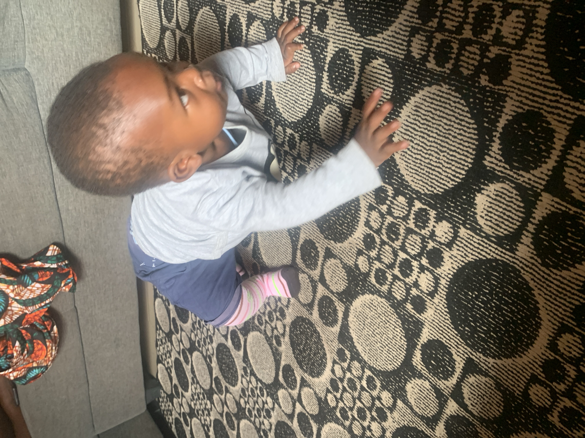 An overhead, slightly angled shot captures a baby crawling on a carpet in Sabon Tasha, Nigeria. The baby, a dark-skinned infant, wears a light blue long-sleeved shirt and dark blue shorts. One leg is visible, clad in a striped pink and white sock with a dark brown toe. The carpet has a repeating pattern of dark circles and geometric shapes against a lighter background. In the background, a portion of a grey sofa is visible, with a folded piece of fabric featuring a vibrant, multicolored African print draped over it. The lighting suggests an indoor setting with ambient light.