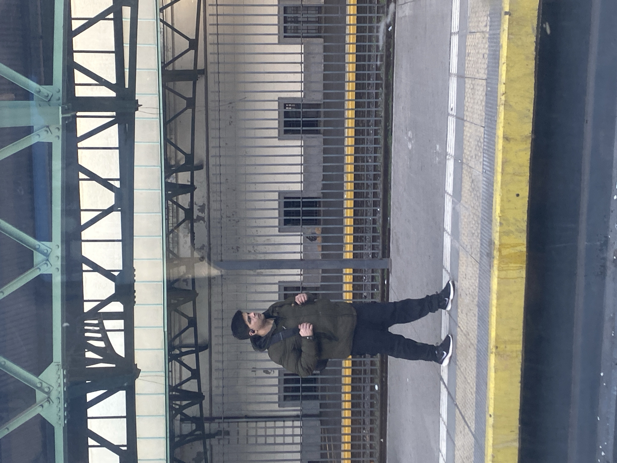 A man stands on a subway platform, wearing a dark beanie, olive green jacket, dark pants, and black and white sneakers. He is facing left and slightly upwards. He has a backpack slung over his shoulder. To his left is a metal fence separating the platform from a building with square window openings. Beyond the fence is a yellow safety line on the edge of the platform. Above and to the left of the man is a green metal structure. The image appears to be taken from a slightly elevated perspective. It is difficult to ascertain the exact location in Buenos Aires, Argentina, without further details about the subway system.