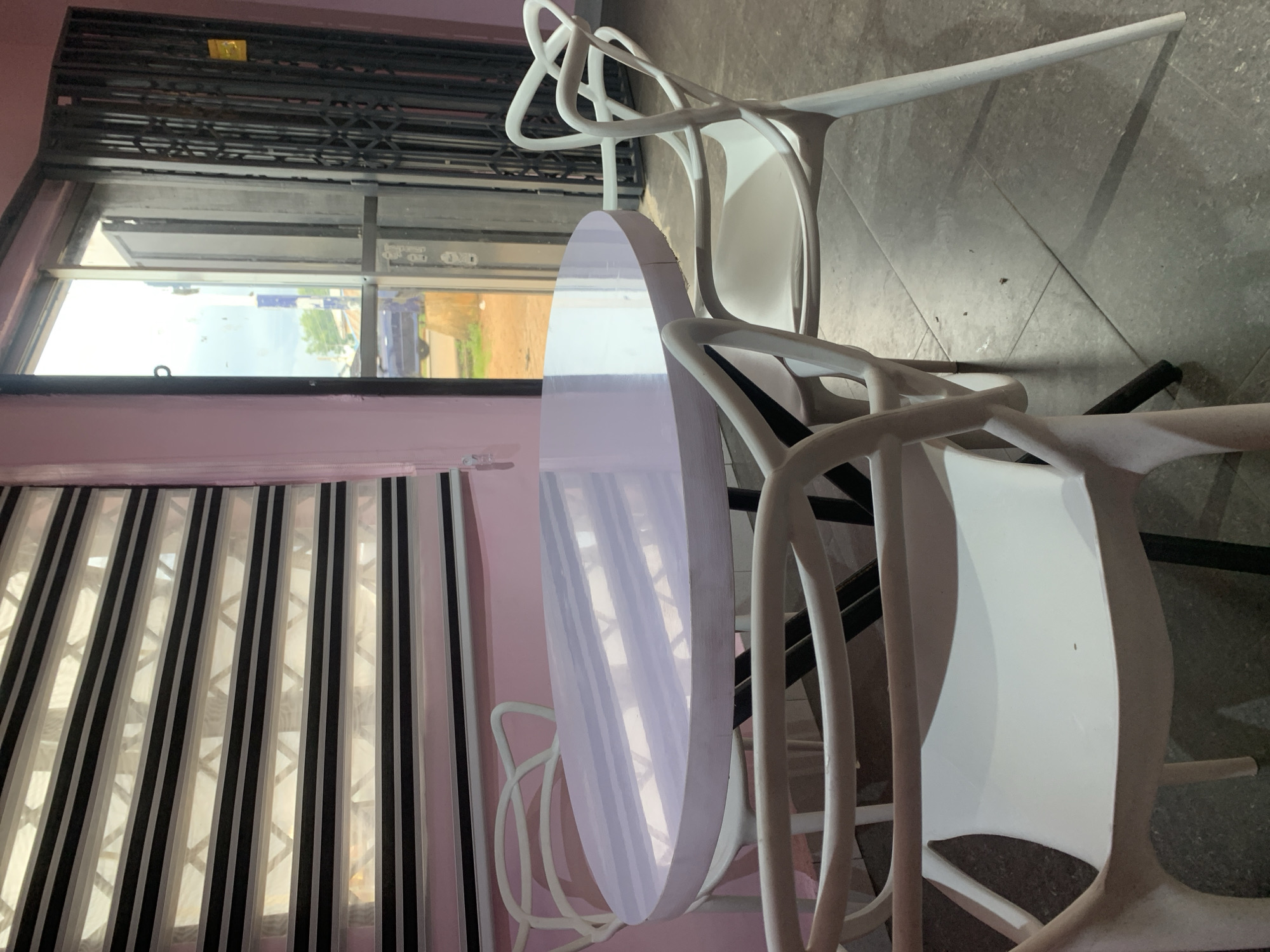 In Jos, Nigeria, a round, light purple table is set with four white chairs inside a room. The table has a striped pattern from sunlight reflecting off its surface. The chairs have a modern design with curved backs and legs. Behind the table, a window with horizontal blinds reveals an outdoor scene with a street and a vehicle. To the right of the window is a metal security gate. The walls of the room are painted pink, and the floor is tiled in gray.