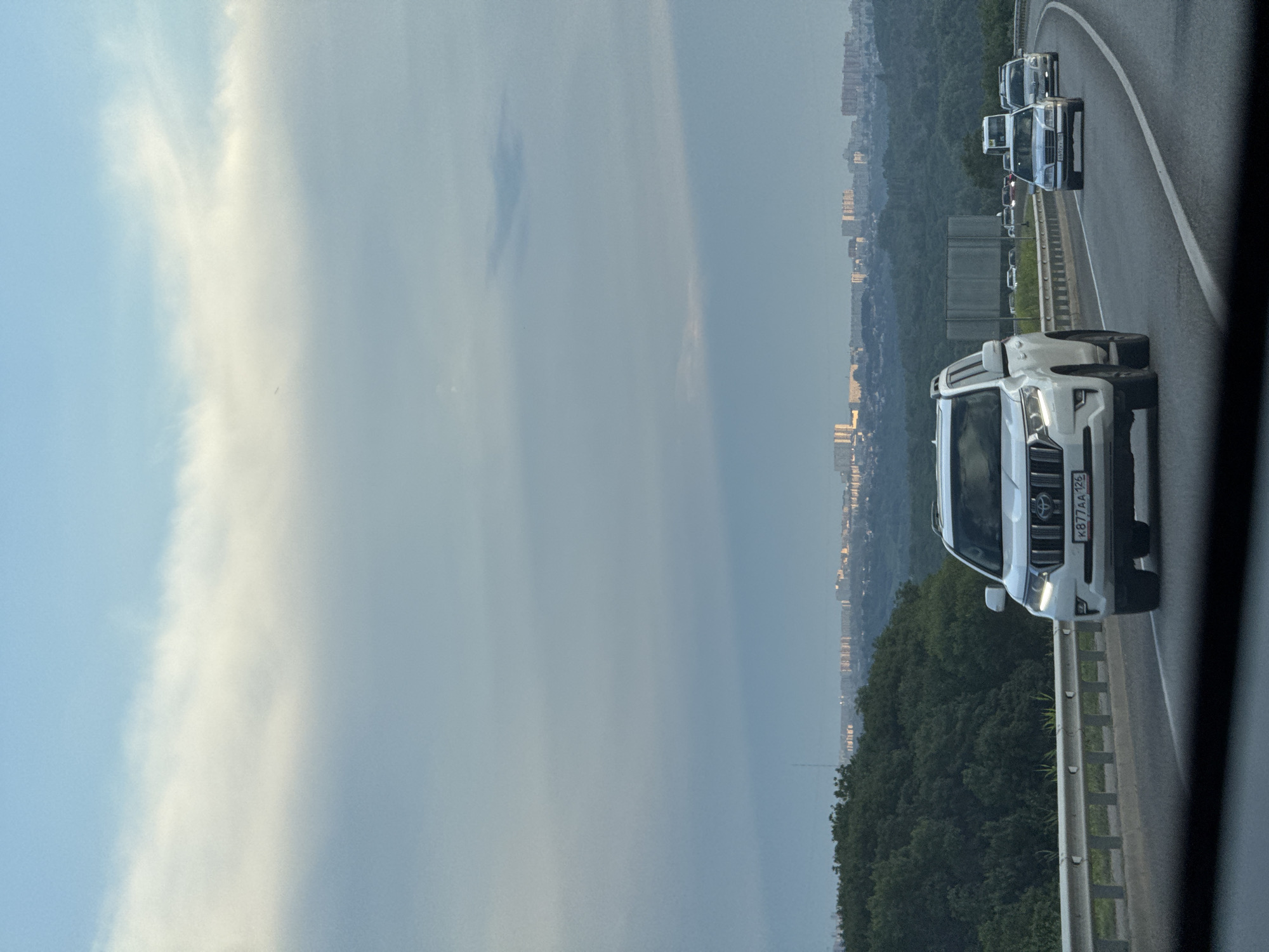 This media file depicts a view from a vehicle traveling on a highway. The primary subjects are several cars on the road, including a white SUV in the foreground with a license plate that reads "K877AA 126". The scene also includes a city skyline in the distance, suggesting an urban or semi-urban setting. Trees line the side of the highway, and the sky is filled with clouds, indicating overcast weather. The time of day appears to be late afternoon or early evening, given the soft, diffused light. The overall mood is neutral, focusing on the transportation aspect of the scene. The location context provided is Novomar'evskaya, Russia.