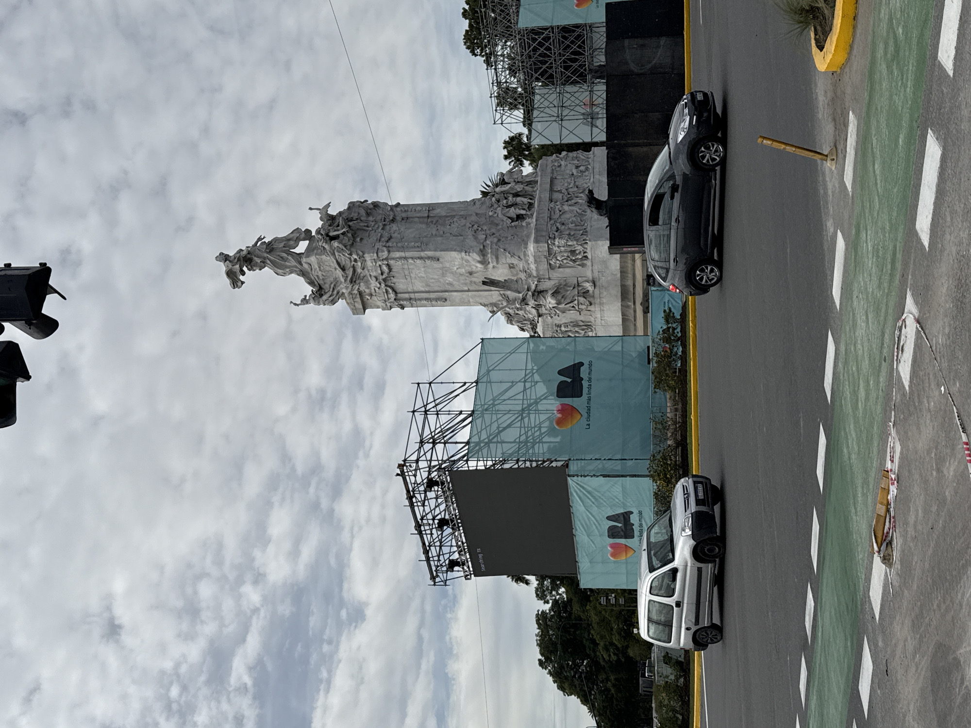 The image depicts the Monument to the Carta Magna and Four Regions of Argentina in Buenos Aires, Argentina. The monument stands tall against a cloudy sky. Scaffolding and banners surround the base of the monument. Two cars are parked along a green and grey road with yellow borders. One car is dark colored, the other is silver. A traffic signal is partially visible in the lower left corner of the image.
