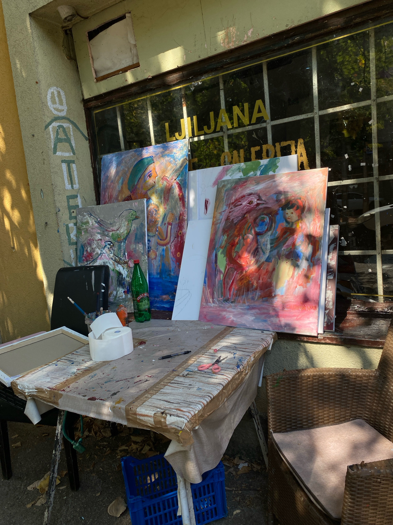 The image depicts an outdoor art studio setting in Cetinje, Montenegro. Several paintings are propped up against a building with a window displaying the word "LJUBLJANA" in yellow lettering. A table covered with a paint-splattered cloth is in the foreground, holding art supplies such as brushes, a roll of toilet paper, a bottle of green liquid, and a pair of pink scissors. Two blue plastic crates are visible beneath the table. A wicker chair is situated to the right of the table. The building's facade is painted in a light yellow and green, with some graffiti present on the wall. The ground is paved, and fallen leaves are scattered around. The scene is illuminated by sunlight, casting shadows.