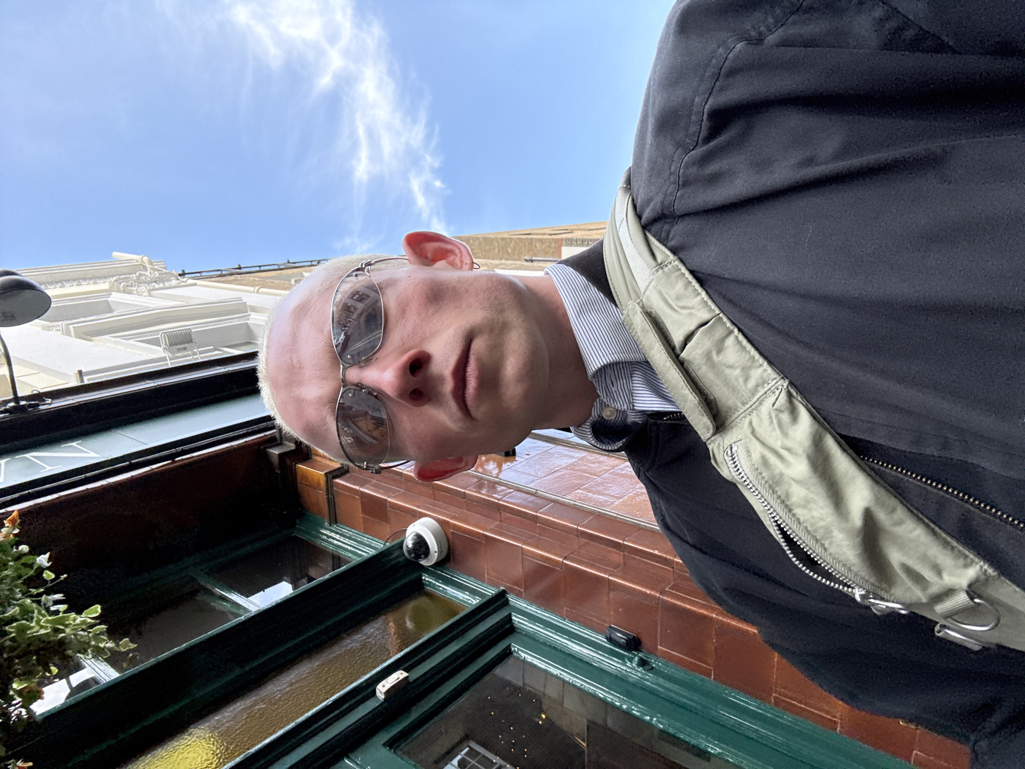 The image is a rotated, low-angle self-portrait taken outdoors on a sunny day in London, United Kingdom. The frame is dominated by the upper torso and head of a person with fair skin, very short light blonde or white hair, and mirrored sunglasses. The person wears a blue and white striped shirt under a dark (likely black) jacket, with a light olive green shoulder bag strap visible across their front. Their expression is neutral.
Behind and above the person, a clear blue sky with sparse white wispy clouds is visible. To the left, a multi-story light-colored building with ornate white architectural detailing, including cornices and windows, ascends towards the sky. Below the person's chin and extending downwards, a section of a building facade features dark green window frames and panelling. Above these windows, a reddish-brown tiled or brick band runs horizontally, with a white dome-shaped security camera mounted on it. Partial green foliage is seen on the far left of the lower building section.
Visible text on the dark green panel of the building includes a clear capital 'N' and incomplete letters, likely part of a sign. The bright daylight indicates it is daytime with clear weather conditions.