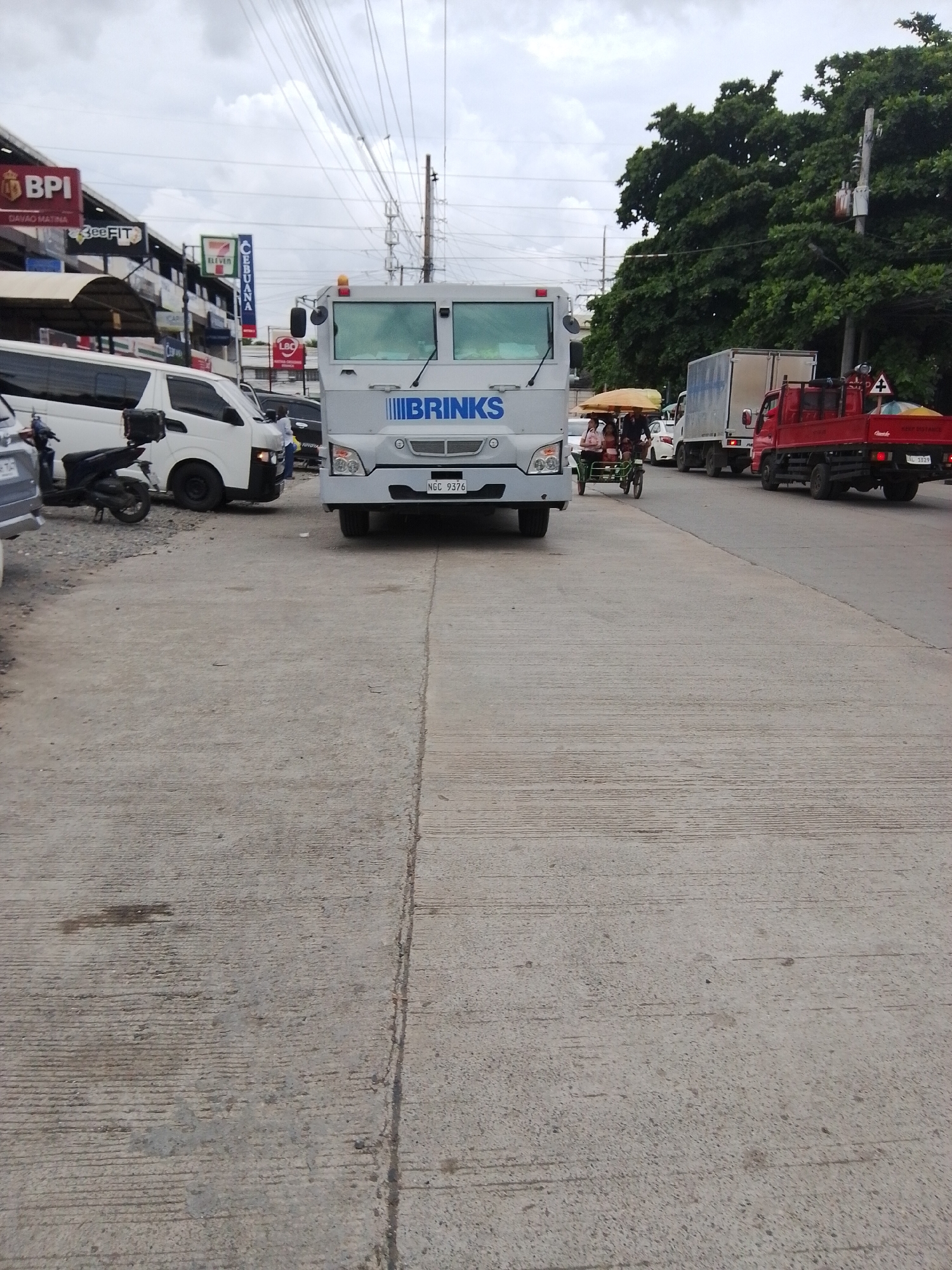 A Brinks armored truck is parked on a street in Davao City, Philippines, with a white van and a red truck also visible. The sky is overcast, and there are trees and power lines in the background. The street is made of concrete, and there are some oil stains and cracks. The Brinks truck has the company logo on the front, and its license plate reads "NGC 9376". A motorcycle is parked next to the white van. There are also some people on a pedicab in the middle of the frame. Businesses like BPI and 7-Eleven are visible in the background.
