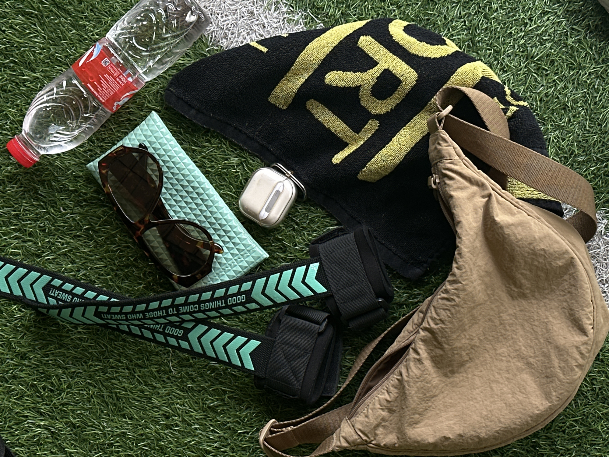 A flat lay of workout accessories is arranged on a green artificial turf surface, possibly a sports field in North Kuta, Indonesia. The items include a light brown, crescent-shaped crossbody bag with a zippered opening and a single strap. Adjacent to the bag are two black resistance bands with turquoise chevron patterns and the text "GOOD THINGS COME TO THOSE WHO SWEAT!". A black towel with a large yellow graphic is also present, along with a pair of tortoiseshell sunglasses resting on a light blue textured mat. A clear plastic water bottle with a red cap and a white label is positioned towards the top right of the frame. A white wireless earbud case is placed on the black towel. The lighting suggests an outdoor setting, possibly during the day. There are no people or discernible actions depicted in the image, focusing solely on the still arrangement of objects.