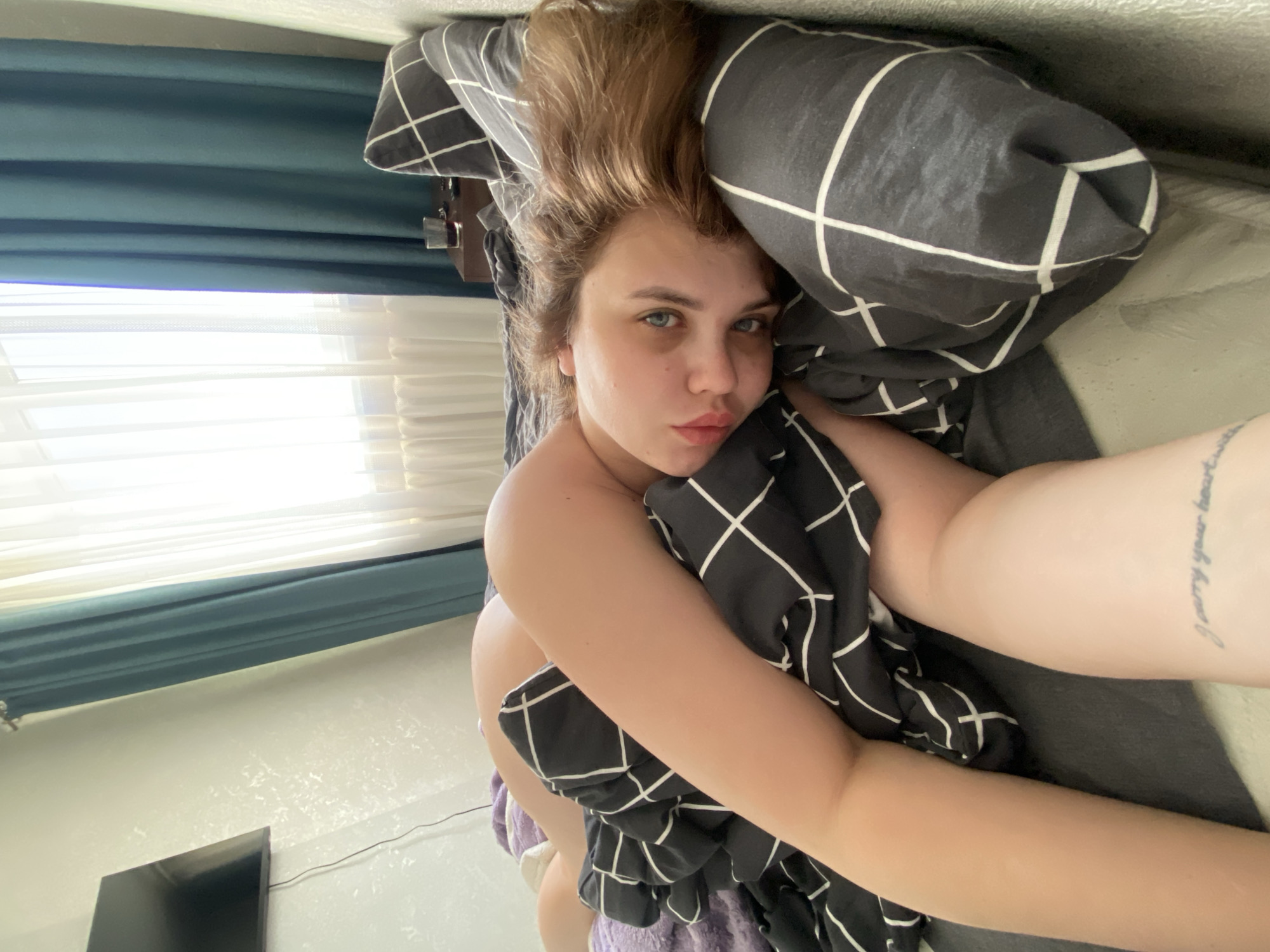 A young woman is captured in a close-up selfie, lying in what appears to be a bed in an indoor setting. Her head is resting on a dark gray pillow with a white grid pattern, and she is partially covered by a matching blanket or duvet, wrapped around her arms and torso. She has light brown or auburn hair that is slightly disheveled, and striking blue eyes. Her lips are slightly pursed in a pouting or kissing expression as she looks directly into the camera.
The background reveals a bright window with sheer white blinds, suggesting it is daytime, possibly morning, given the soft, bright light. Dark teal curtains are visible above the blinds. To the left of her head, a small wooden shelf or nightstand with a few indistinct items is visible. On the lower left side of the frame, a dark flat-screen television is mounted on a wall with a slightly textured, light-colored surface.
A tattoo with script text is visible on her left forearm, reading "I carry your heart". The overall atmosphere is relaxed and intimate. The image is set in Chernushka, Russia.
