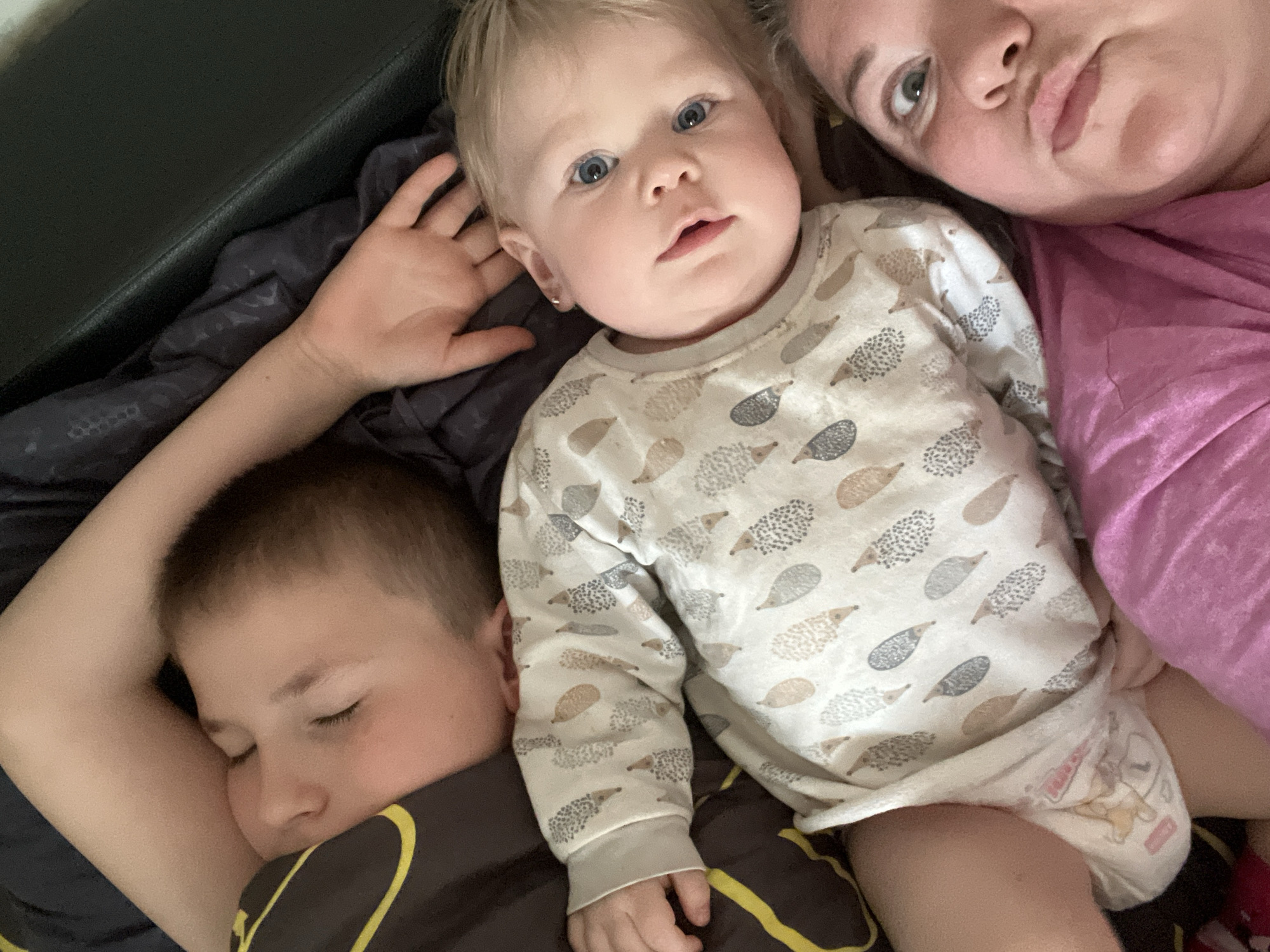 This close-up, high-angle image captures an intimate family moment indoors in Фоки, Russia, likely on a bed or couch. Three individuals are visible: a woman, a baby, and an older child.
In the center, a fair-haired baby with bright blue eyes looks directly at the camera, exhibiting a curious or neutral expression. The baby wears a light-colored, long-sleeved onesie patterned with grey, brown, and beige hedgehogs. A small stud earring is visible in the baby's left ear. The baby's left hand is partially seen behind its head, resting on dark bedding, while its right hand rests on the older child. A white diaper with an illustrated character (possibly "DanDan") and some visible text is worn underneath the onesie.
To the bottom left, an older child with short brown hair is sleeping soundly, their eyes closed. They are partially covered by dark bedding and wear a dark top with yellow accents. Their left arm is visible, bent under their head.
In the upper right, part of a woman's face and upper body are visible. She appears to be looking towards the baby or the camera, with a neutral or slightly tired expression. She is wearing a pink top.
The setting appears comfortable and relaxed, possibly during a morning or evening snuggle. The lighting is soft, consistent with an indoor environment. The dark bedding and a black headboard or furniture piece are also visible in the background.