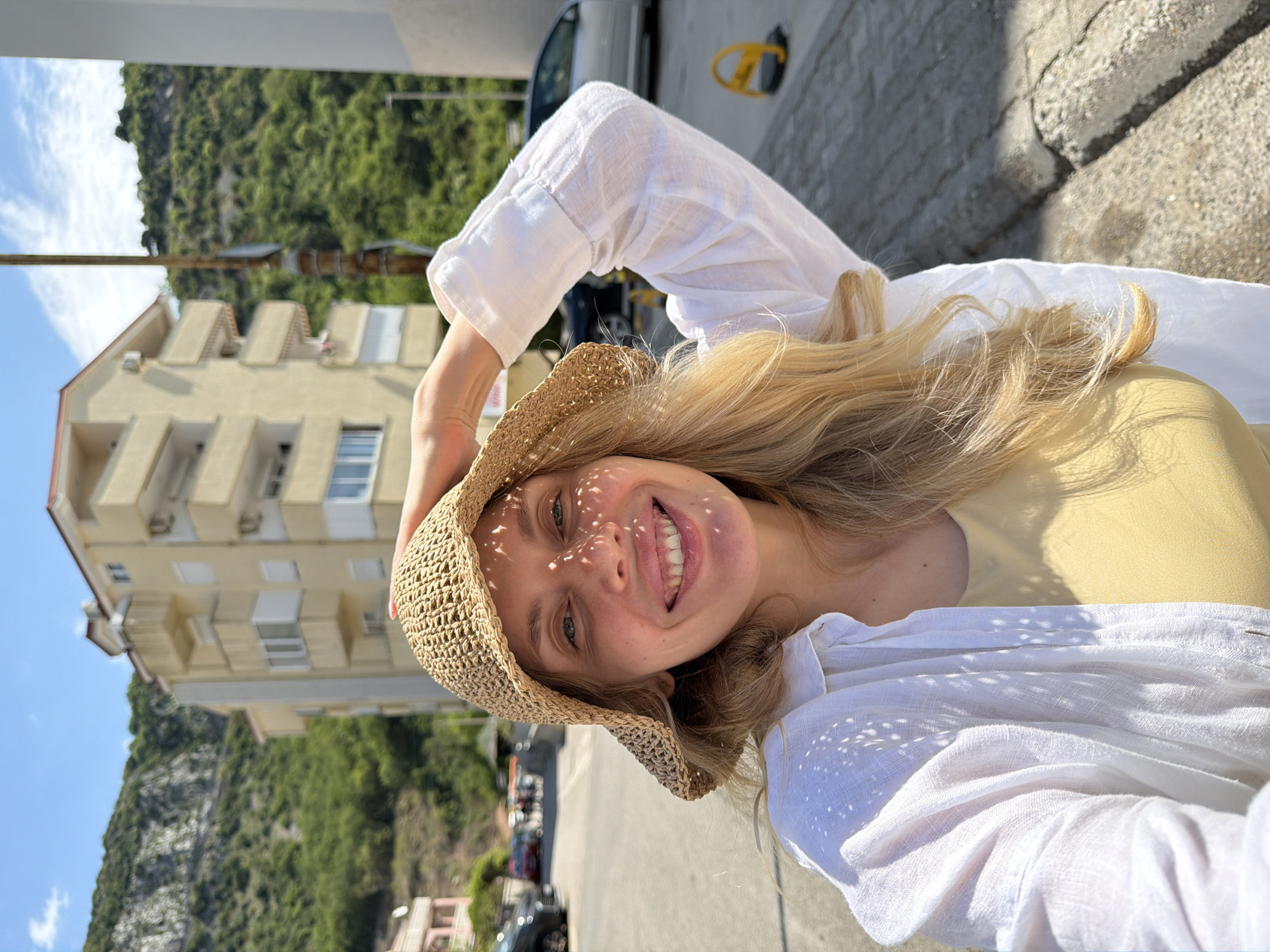 The image is an outdoor selfie featuring a smiling young woman against a sunny, mountainous backdrop. The woman, with long blonde hair, wears a light-colored top under a white, long-sleeved, possibly linen, shirt. She is also wearing a woven straw hat, through which sunlight filters, casting a distinctive dappled pattern of light and shadow across her face and shirt. Her wide smile conveys happiness and relaxation.
The setting appears to be a street or sidewalk in a coastal town. In the background, multi-story, light-colored apartment buildings with balconies and shutters are visible, consistent with Mediterranean architecture. Behind the buildings, a large, densely vegetated green mountain rises prominently under a clear blue sky with sparse white clouds. The ground is a paved surface, showing some cracks. Strong sunlight and clear skies indicate it is daytime with pleasant weather. A partial view of a vehicle and a yellow street sign or parking barrier can be seen in the background. The overall scene, with its architectural style and mountainous coastal landscape, is consistent with the provided location context of Budva, Montenegro. No visible text is present in the image.