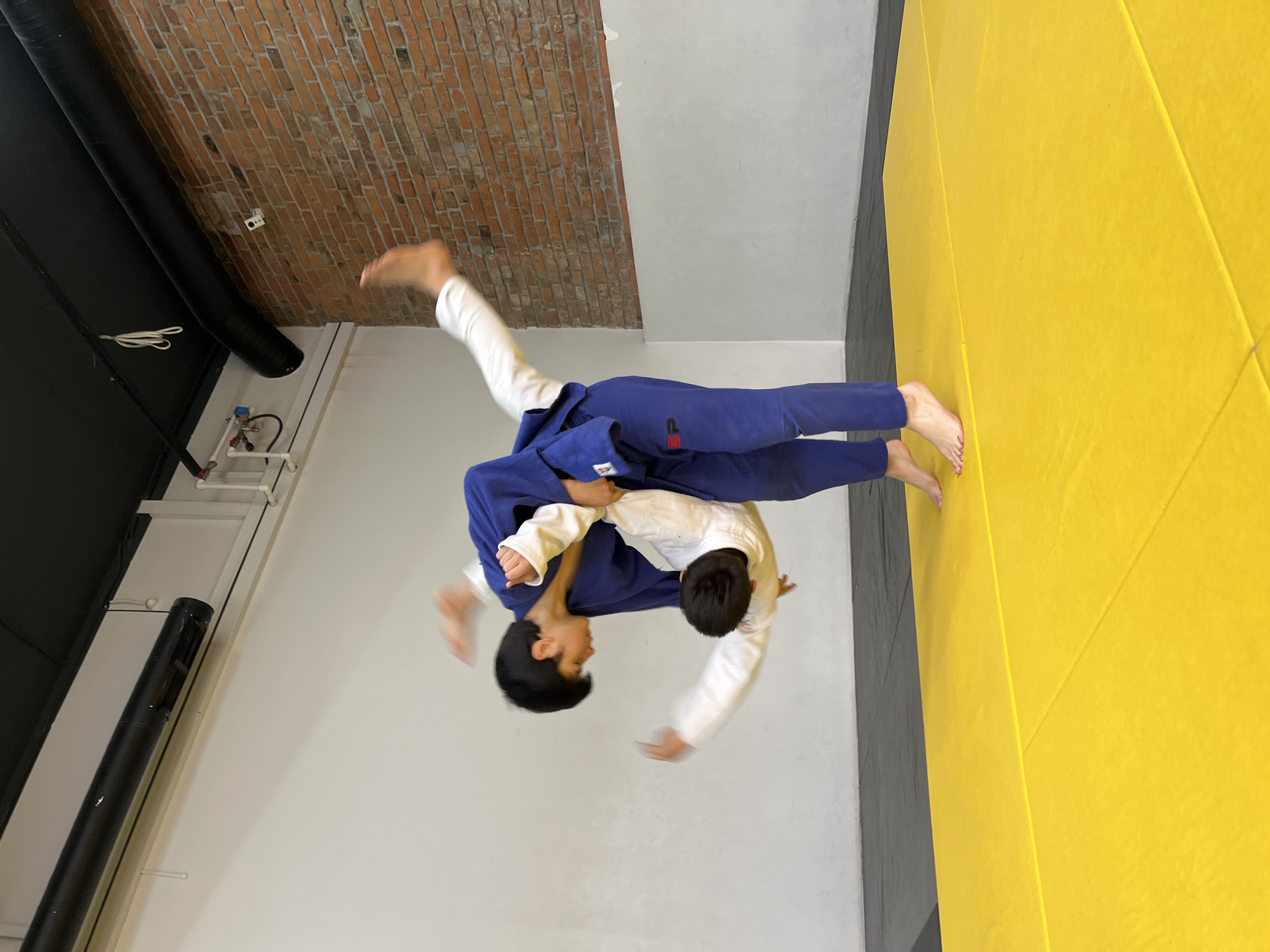 Two individuals are engaged in a martial arts activity within an indoor training facility. One individual, wearing a blue uniform, is executing a throwing technique on another individual wearing a white uniform. They are positioned above a yellow mat. The location appears to be a gymnasium or specialized training area, possibly in the city of Krasnoyarsk, Russia. The background includes a brick wall, white walls, and what seems to be ventilation or plumbing elements on the ceiling.