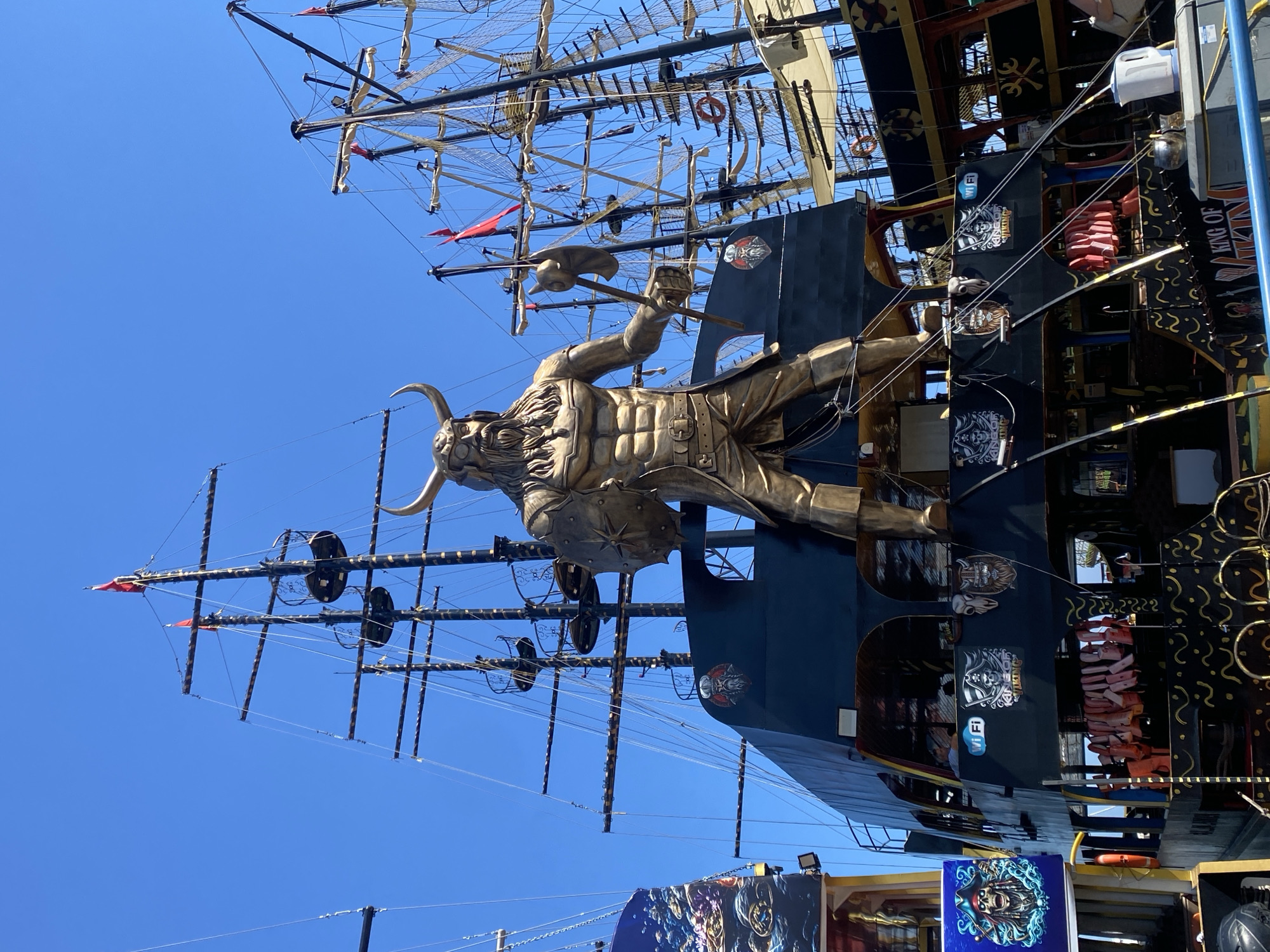 The image shows a pirate ship-themed tourist attraction in Alanya, Turkey. The foreground features a large, ornate, golden-colored statue of a horned, mythological figure with a beard, wearing armor and holding a weapon. The statue is positioned on the side of a dark-colored ship with intricate gold detailing, including decorative scrollwork and skull-themed emblems. The ship's mast extends upwards, with visible rigging, sails, and flags. The sky is clear and blue. The ship appears to be docked, as evidenced by the presence of nearby structures and signage, including a sign with the text "KING OF". Other signs and decorative panels featuring images of pirates and nautical themes are visible. There are life vests stacked in the interior.