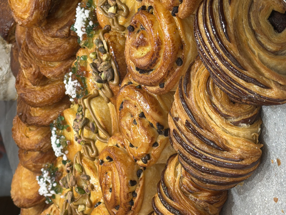A close-up, high-angle view captures a dense, multi-layered display of various French pastries, likely situated within a patisserie or boulangerie in Paris, France. The scene is static, devoid of people or direct action.
In the foreground, several elongated, golden-brown laminated pastries feature prominent dark chocolate stripes across their ridged, flaky surfaces. Immediately behind these, multiple circular, spiral-shaped pastries are visible, characterized by a glossy golden-brown glaze and embedded dark chocolate chips.
Further back, a row of similar spiral pastries presents diverse toppings: some are adorned with green pistachio pieces and a pale green or beige drizzle, while others are topped with a green translucent jelly-like substance and small white sugar pearls.
The background shows additional pastries, including round, layered, golden-brown items with a distinct folded texture, suggesting *kouign-amann* or similar viennoiseries, arranged in dense rows. The pastries are uniformly baked to a golden-brown hue, often with a glossy finish, and are displayed on a light, neutral-toned surface.