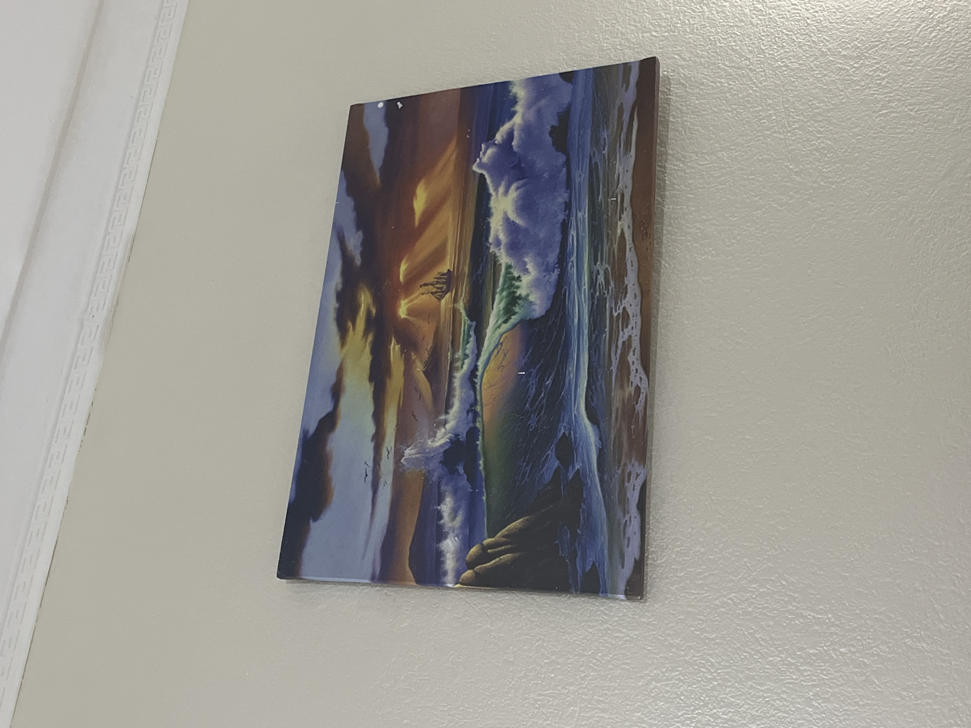 The image shows a framed painting of a seascape with a sunset, a sailboat, and crashing waves. The painting is hung on a textured, light-colored wall, just below a white decorative molding. The scene depicted in the painting features a dramatic sky with shafts of sunlight breaking through dark clouds. A sailing ship is visible in the distance. In the foreground, large waves are breaking against rocks and on a sandy shore. The painting's surface appears glossy. The location is not specified within the image itself, therefore no information about Bashkia Durrës, Albania can be provided.