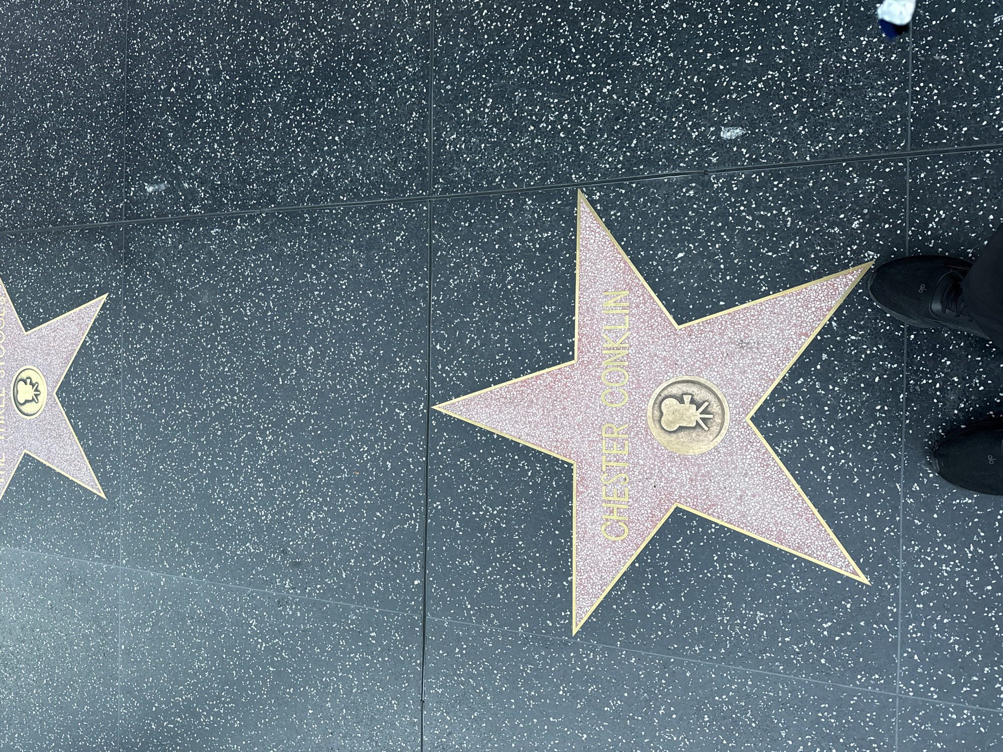 This image captures a ground-level view of two Hollywood Walk of Fame stars embedded in a dark gray, speckled terrazzo sidewalk. The prominent star on the right features the name "CHESTER CONKLIN" in gold capital letters, oriented vertically. Below the name, a gold circular emblem depicts a classic film camera, indicating his contribution to motion pictures. This star is a five-pointed design with a light pinkish-gold color and a golden border.
A person's black shoe is visible in the top right corner, partially covering the top point of Chester Conklin's star. To the left, a portion of another similar star is visible, with only its right edge and some indistinct text showing. The sidewalk has faint grout lines forming a grid pattern. The scene depicts a typical daytime setting on the Hollywood Walk of Fame in Los Angeles, USA. Minor scuff marks and small pieces of debris are scattered across the pavement.