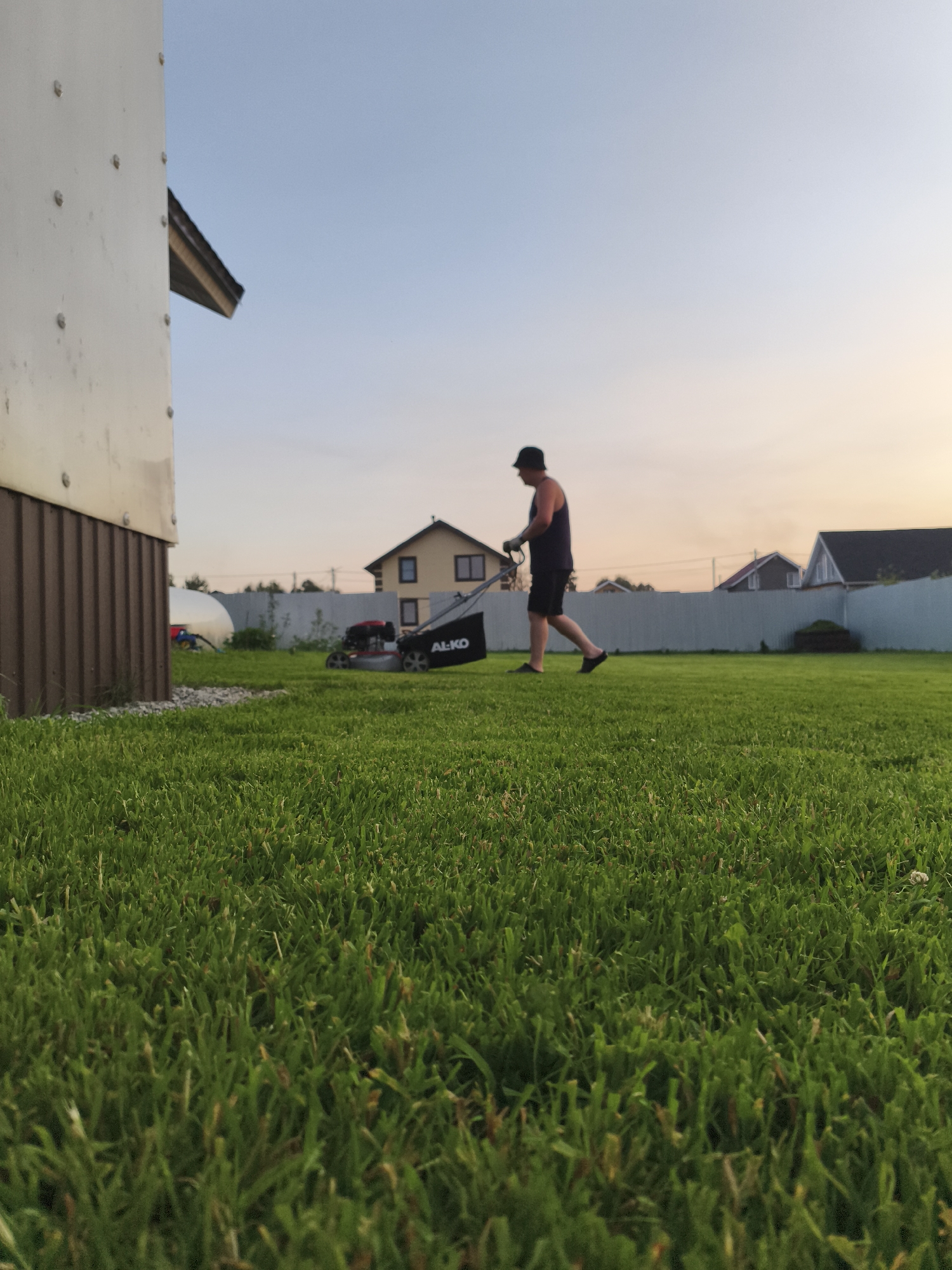The image shows a person mowing a lawn in Русский Вожой, Russia. The person, wearing a dark tank top, black shorts, a black baseball cap, and dark flip-flops, is operating a red and silver lawnmower with a black collection bag that has "AL-KO" printed on it. The lawnmower is on a well-maintained, bright green lawn. In the background, there is a light-colored house with a dark roof, a white fence, and several other houses with similar architectural styles. The sky is a soft gradient of blue and yellow, indicating it is either early morning or late evening. A portion of a building, possibly a shed or garage, is visible on the left side of the frame, with a corrugated metal siding and a light-colored upper section. The foreground is dominated by the lush green grass, with the camera positioned at a low angle.