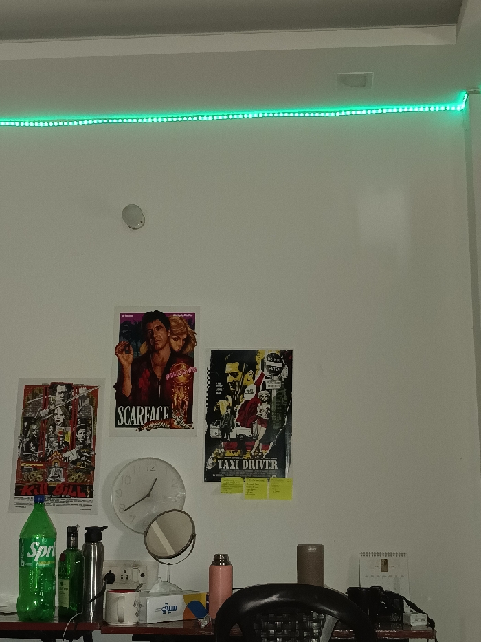 The image shows the interior of a room in Ghaziabad, India, located at 28.682 latitude and 77.41 longitude. The room has a white wall with a green LED light strip mounted on the ceiling. There are three movie posters on the wall: a "Scarface" poster featuring Al Pacino and a woman, a "Taxi Driver" poster featuring Robert De Niro and a woman, and a poster for the movie "Kill Bill" featuring Uma Thurman. There is a clock on the wall, a round mirror on a stand, a bottle of green soda labeled "Sprite" on a table, a silver water bottle, a pink thermos, a brown ceramic mug, a box of facial tissues labeled "Sawan," and a black wicker-backed chair. The room has a desk with a black surface and a black camera on top of it. The camera is positioned in front of the black chair. The room appears to be a bedroom or office space.