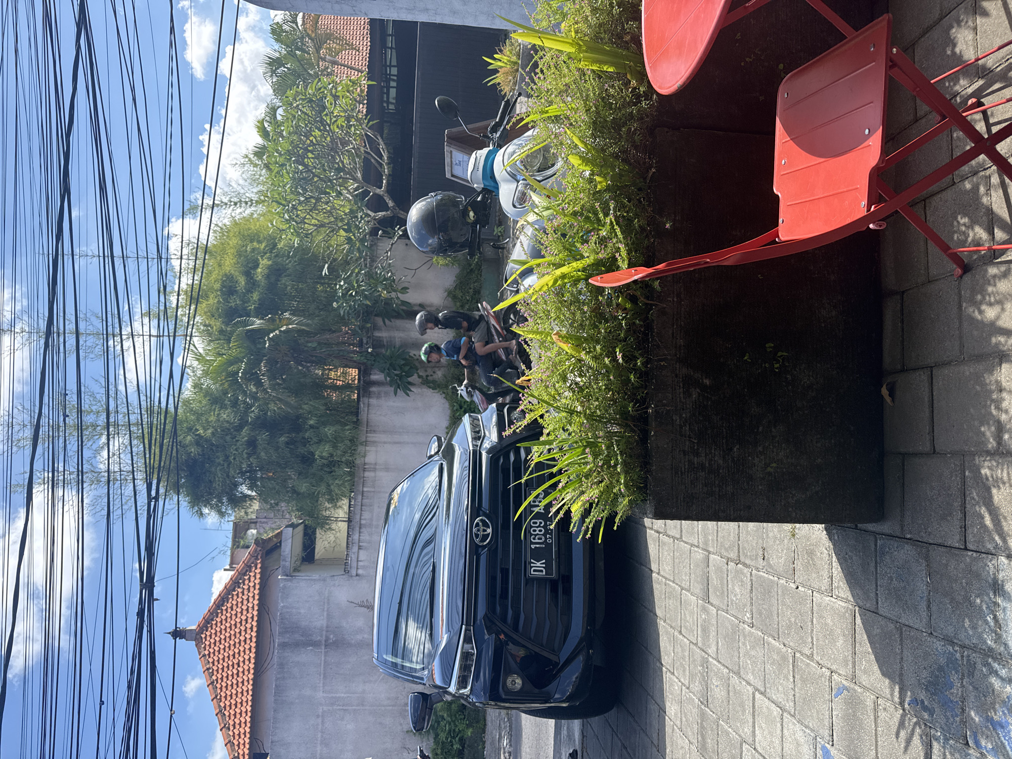 This image depicts an outdoor street or pathway scene in North Kuta, Indonesia, under bright daylight. In the mid-ground, a dark-colored Toyota SUV is prominently parked on a grey paved surface, displaying the Indonesian license plate "DK 1689 ABS" with "07 27" below it, indicating a Bali registration. Behind the SUV, several motorbikes are visible, including a white Honda scooter, with two individuals standing nearby. One person wears a black helmet, and the other a green helmet, suggesting they are either preparing to ride or have recently arrived.
To the right in the foreground, a section of an outdoor seating area features two bright red metal chairs and part of a matching table. Adjacent to this seating is a large, dark planter overflowing with lush green foliage, including ferns and various flowering plants. The ground is covered with grey interlocking pavers, and distinct shadows indicate strong sunlight, suggesting a clear day, likely mid-morning or afternoon.
In the background, buildings with traditional terracotta-tiled roofs are visible, interspersed with abundant tropical greenery, including tall bamboo and palm trees. The bright blue sky, dotted with white clouds, is crisscrossed by numerous black utility wires that stretch across the upper portion of the frame. A small sign on the white Honda scooter has some visible text, partially obscured, which may say "Thank You."