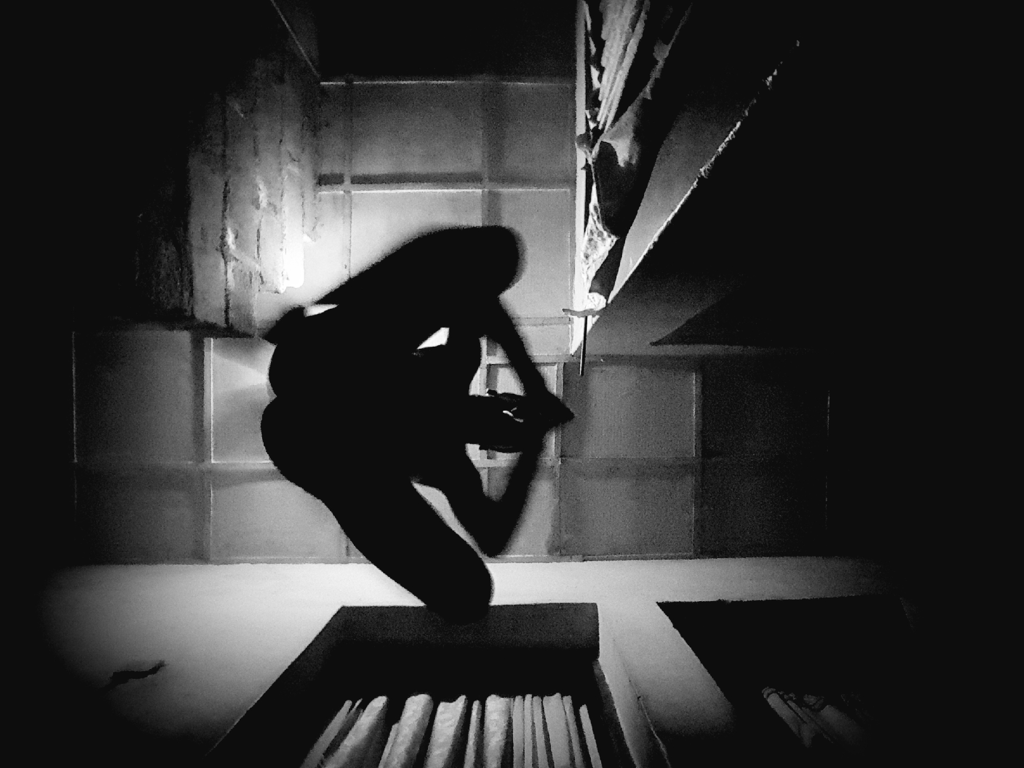 The image is a black and white, high-angle shot of a person silhouetted against a textured background. The person is in a seated or squatting position with their legs bent and spread wide, and their arms are raised, hands clasped behind their head. The lighting creates strong shadows, obscuring fine details of the subject. The environment appears to be an interior space with a grid-like pattern on the walls or floor, and some stacked objects are visible to the right. There are no recognizable landmarks or elements that place the scene specifically in Narayi, Nigeria. The overall impression is one of abstract form and shadow play.