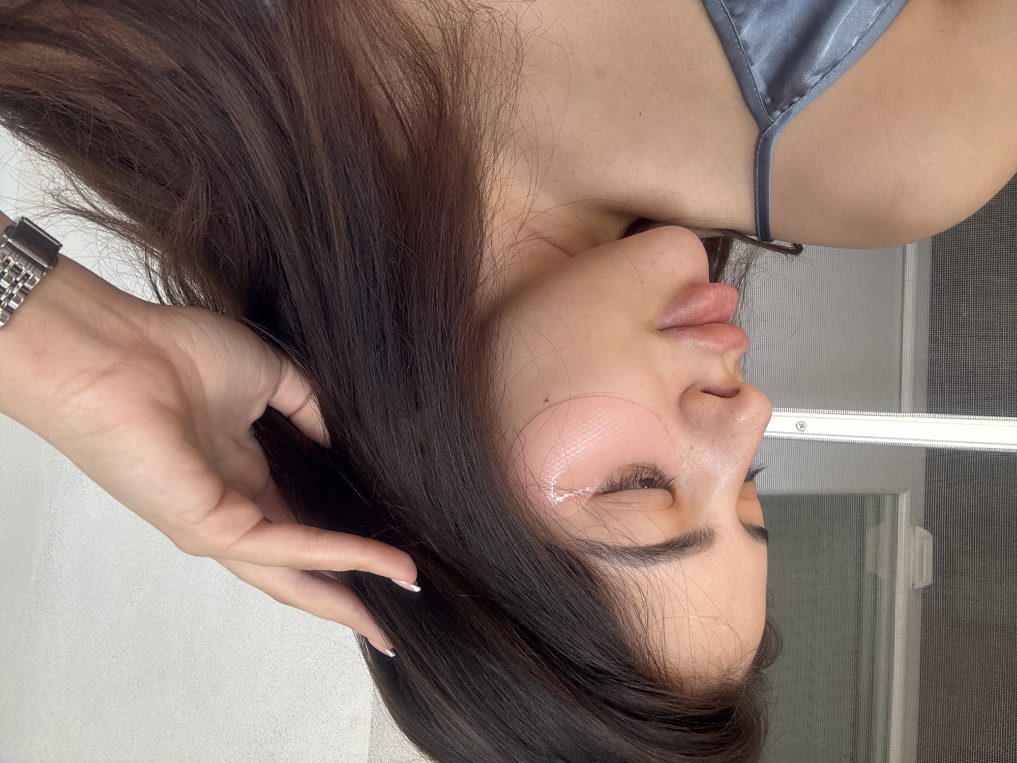 The image captures a close-up portrait of a woman, seen from a side profile, against a simple indoor background. She appears to be reclining or lying down, with her head resting on her side, eyes closed or partially closed, suggesting a moment of relaxation or rest. Her long, dark brown hair cascades around her face and shoulder.
She is wearing two pink hydrogel eye patches, one under each eye, indicating a skincare or beauty treatment. A light blue or grey satin strap of her clothing is visible near her shoulder. On her left wrist, a metallic watch with a link bracelet is noticeable. One of her hands, with manicured nails featuring white tips, is gently placed near her hair on the left side of the frame.
The background consists of a plain white or light-colored wall on the left and a window with a fine mesh screen on the right. A metallic window frame and a visible screw are present in the upper right quadrant. The overall lighting is soft and even, suggesting an indoor setting during the daytime.
The woman's expression is calm and peaceful, reinforcing the idea of relaxation. There is no visible text in the image. The scene takes place in Plovdiv, Bulgaria.