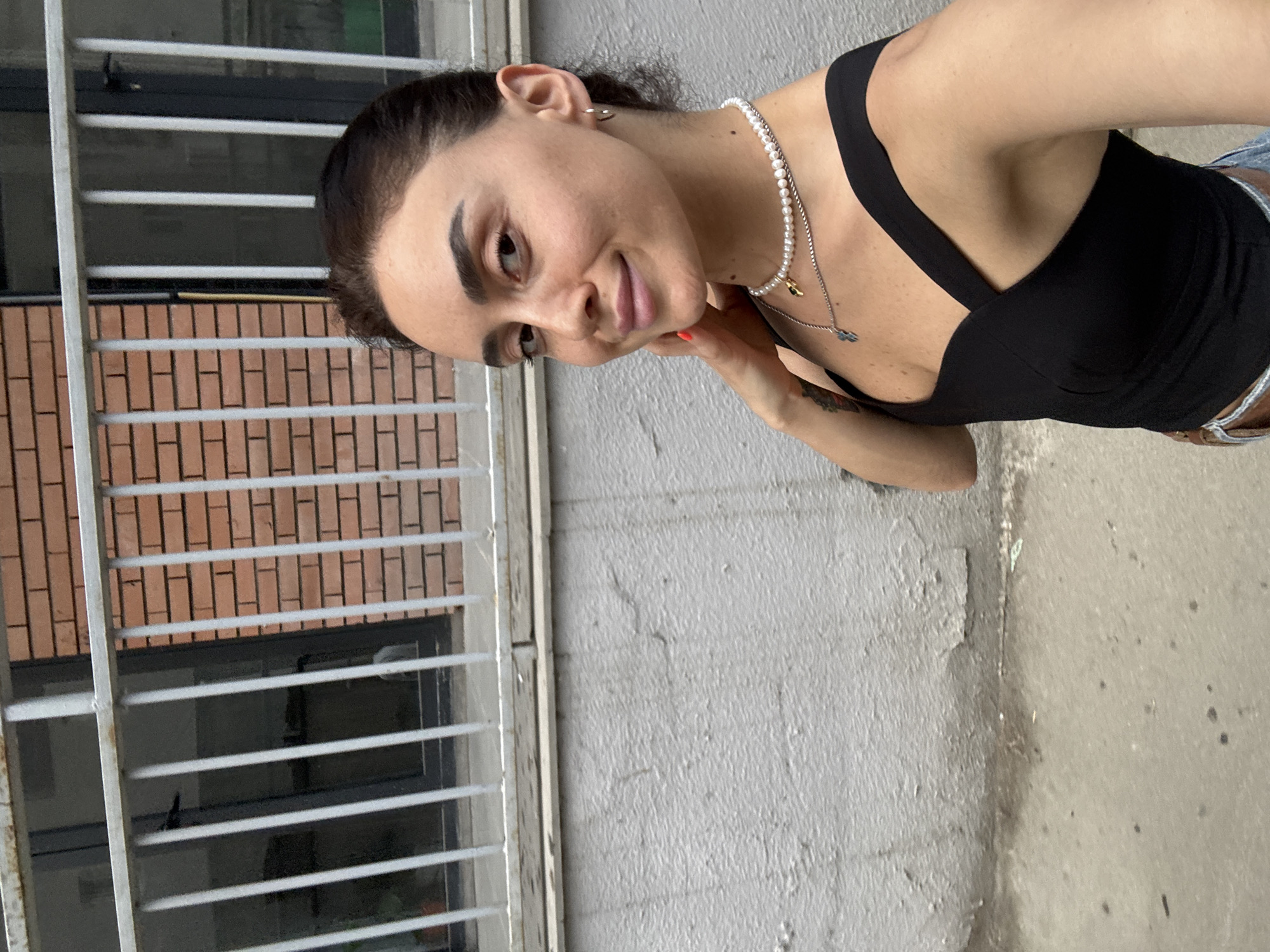 A young woman with dark hair pulled back is leaning her head on her hand, looking towards the camera with a slight smile. She is wearing a black tank top, a pearl necklace, and a silver necklace with a charm. She has a small earring in her left ear. The background shows a brick wall with horizontal white bars in front of it, suggesting a window or a fence. The overall setting appears to be outdoors, with a textured concrete wall on the right side of the image. The lighting suggests it might be daytime, possibly overcast or in the shade. The woman's expression is relaxed and friendly. There is no visible text in the image.