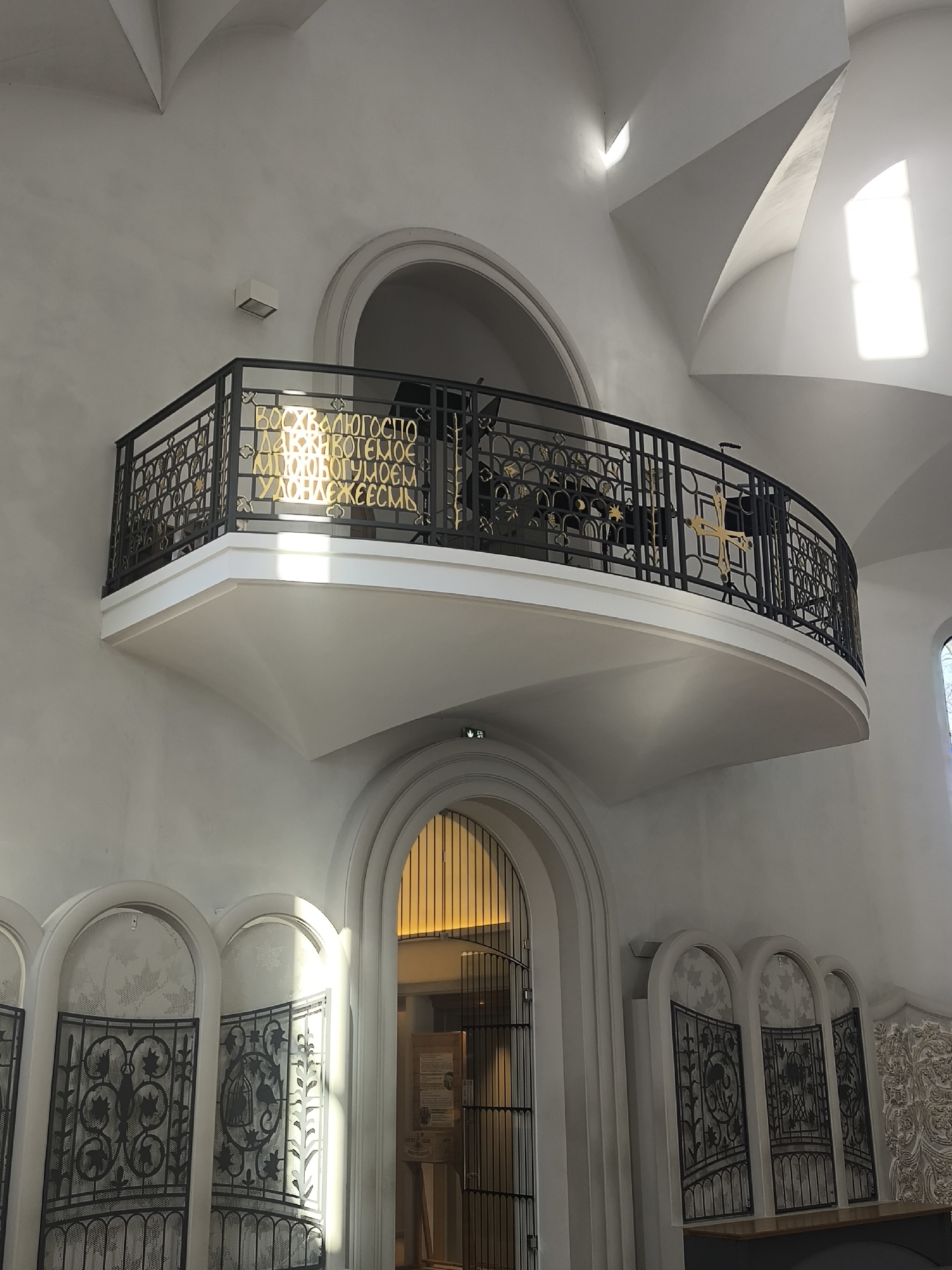 The image depicts an interior space in Strasbourg, France, likely a church or religious building, showcasing a raised balcony with a decorative metal railing. The railing features Cyrillic text in gold lettering. Below the balcony, arched doorways or niches are visible, adorned with decorative metalwork. Sunlight streams through a window in the upper right, illuminating the white walls and highlighting the architectural details. The overall impression is of a serene and elegant space.