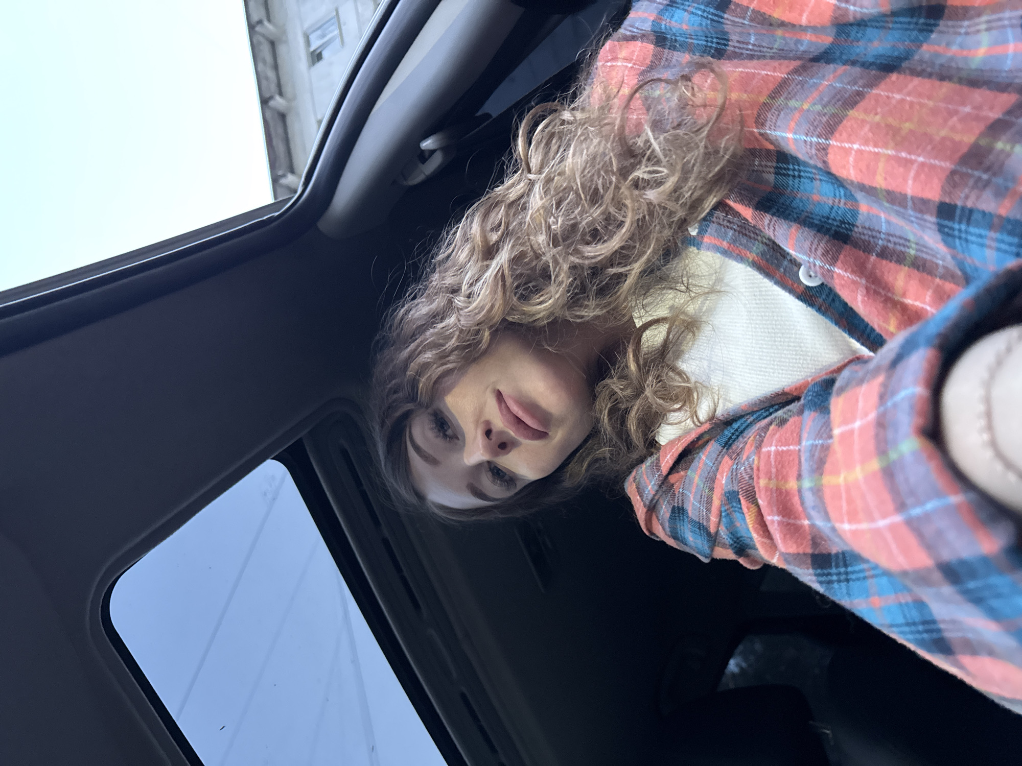 The photograph shows a woman with light brown curly hair wearing a red and blue plaid shirt. She is inside a car, presumably looking upwards at the camera through an open sunroof. The car's interior is dark, and the sky is visible through the sunroof. The architecture of buildings can be seen in the sky. The image was taken in a location in the city of Chaikovsky, Russia.