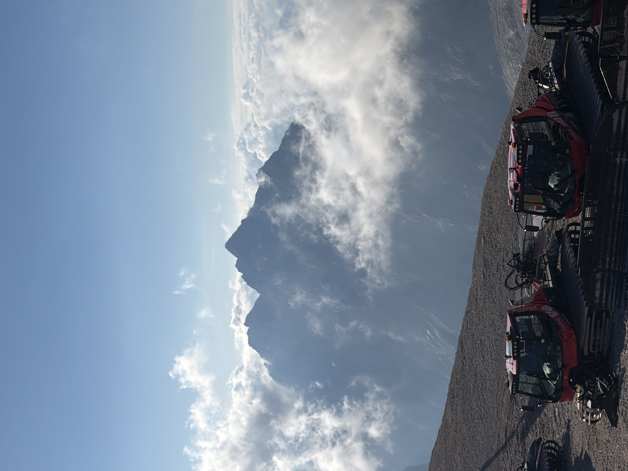 The image depicts a scenic view from a mountain in Garmisch-Partenkirchen, Germany, likely at a ski resort. In the foreground, several red snow groomers are parked on a gravelly slope. These vehicles are designed for maintaining snow surfaces on ski slopes. Behind the snow groomers, a dramatic mountain range emerges from a sea of clouds, with sharp peaks silhouetted against a clear blue sky. The clouds are dense and form a soft, ethereal layer, suggesting a high altitude or a particular weather condition. The overall impression is one of natural grandeur and the presence of specialized equipment for winter sports operations.