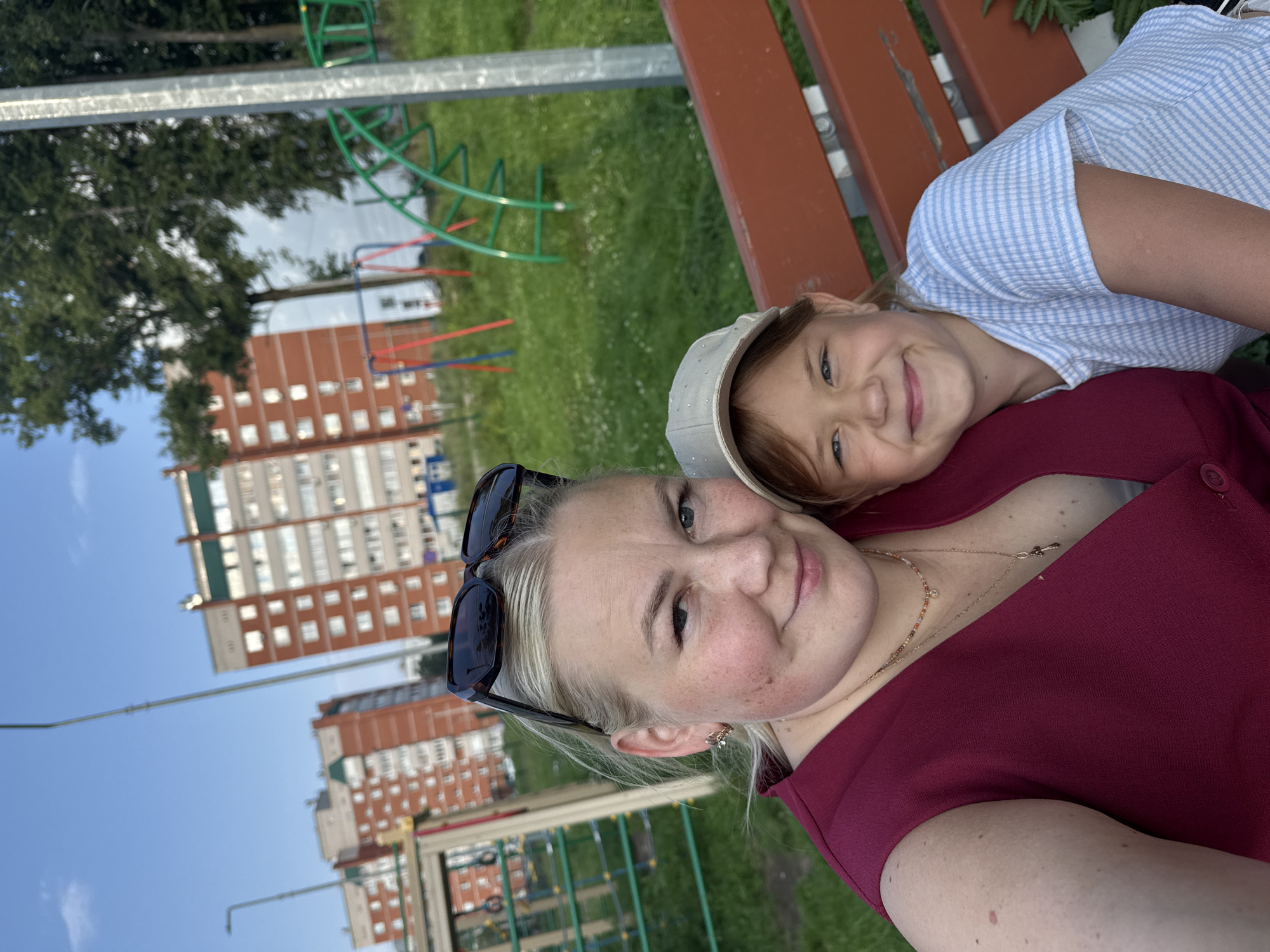 In Chaikovsky, Russia, a woman and a young girl are seated on an orange park bench, taking a selfie. The woman, on the left, has blonde hair pulled back and is wearing sunglasses on her head, a maroon V-neck top, and two necklaces. The girl, on the right, wears a light beige baseball cap and a blue and white striped t-shirt. Behind them, a playground with green and red structures is visible, along with several multi-story brick residential buildings with white-framed windows. The scene is set outdoors with a clear blue sky and green grass.