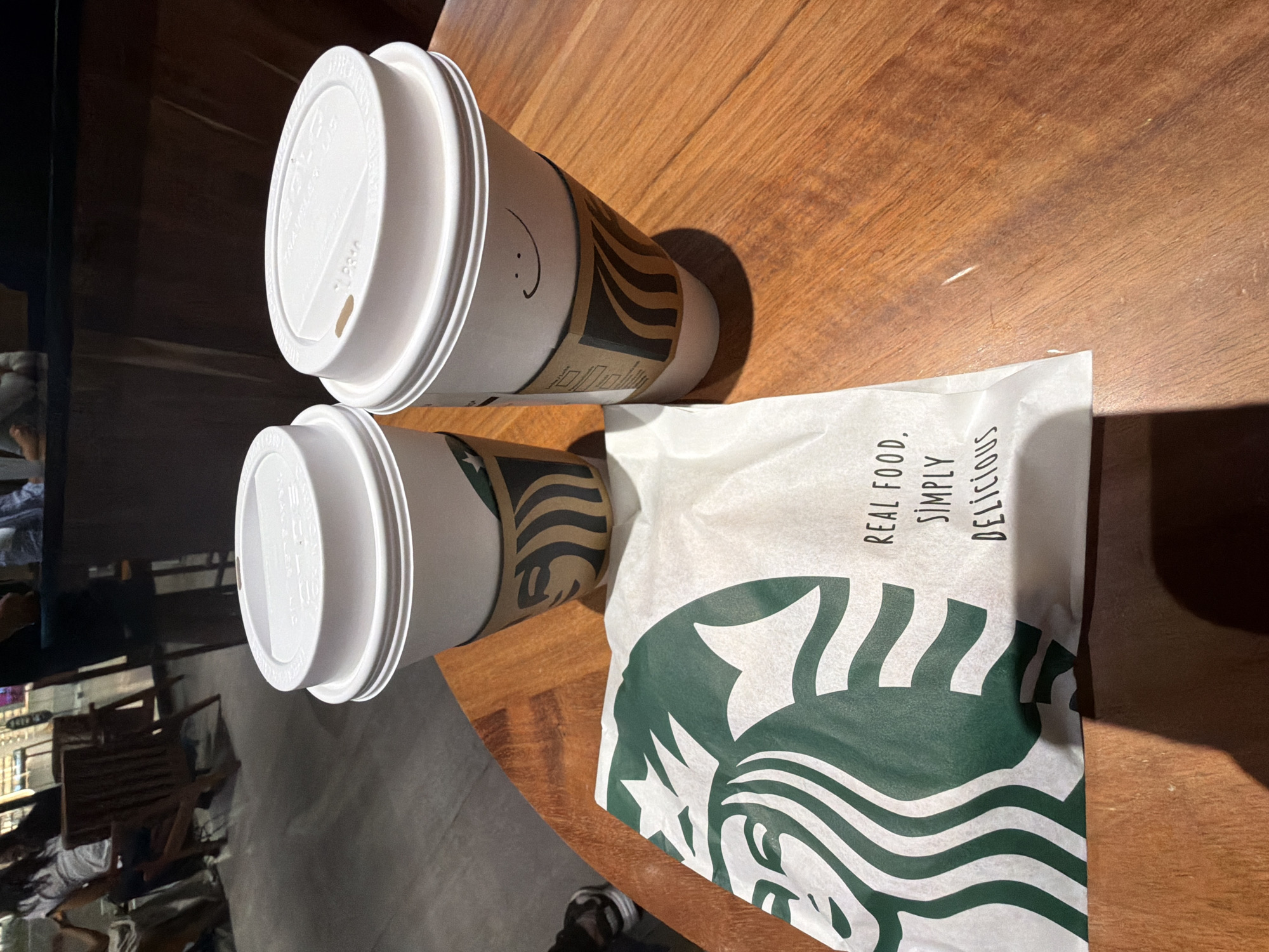 Two cups of Starbucks coffee with white lids, one with a smiley face drawn on it, and a Starbucks bag are on a wooden table. The cups have Starbucks branded sleeves. The bag displays the Starbucks logo prominently along with the text "REAL FOOD. SIMPLY DELICIOUS." The background shows a blurred interior possibly inside the Starbucks location, featuring furniture and people. The location in the city is unknown.