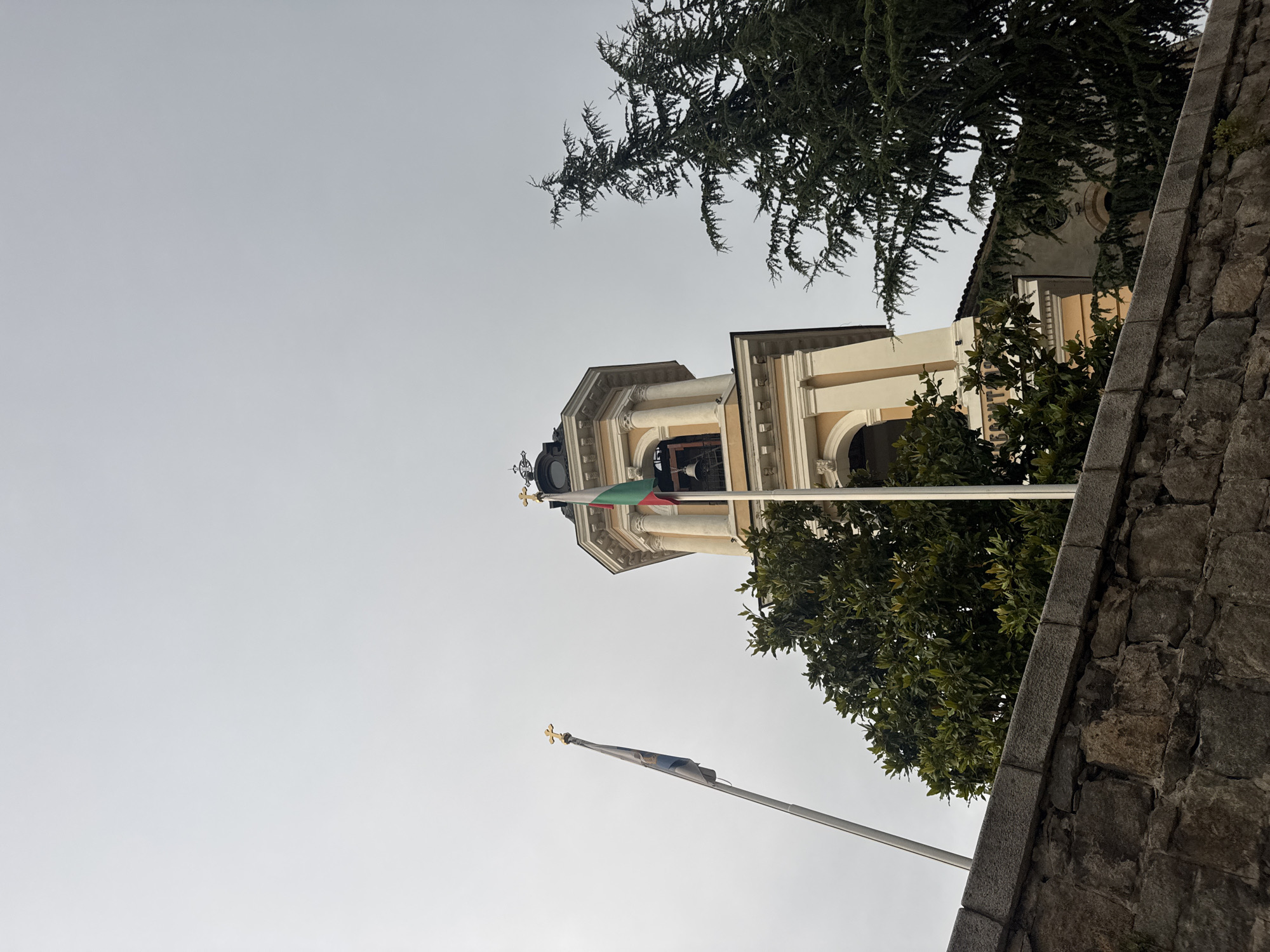 This is a low-angle outdoor, medium shot of a church bell tower in Plovdiv, Bulgaria. The tower is made of light-colored stone with yellow accents and has a black, domed roof topped with a cross. Two flags are visible on flagpoles in front of the tower. One flag is blue and white with a golden cross at the top. The other flag is red, white, and green, and is also topped with a golden cross. A large bell is visible in the open archway of the tower. There are two trees in the foreground, one a large evergreen and the other a tree with dense green leaves. A stone wall runs along the bottom of the image. The sky is overcast and grey.
