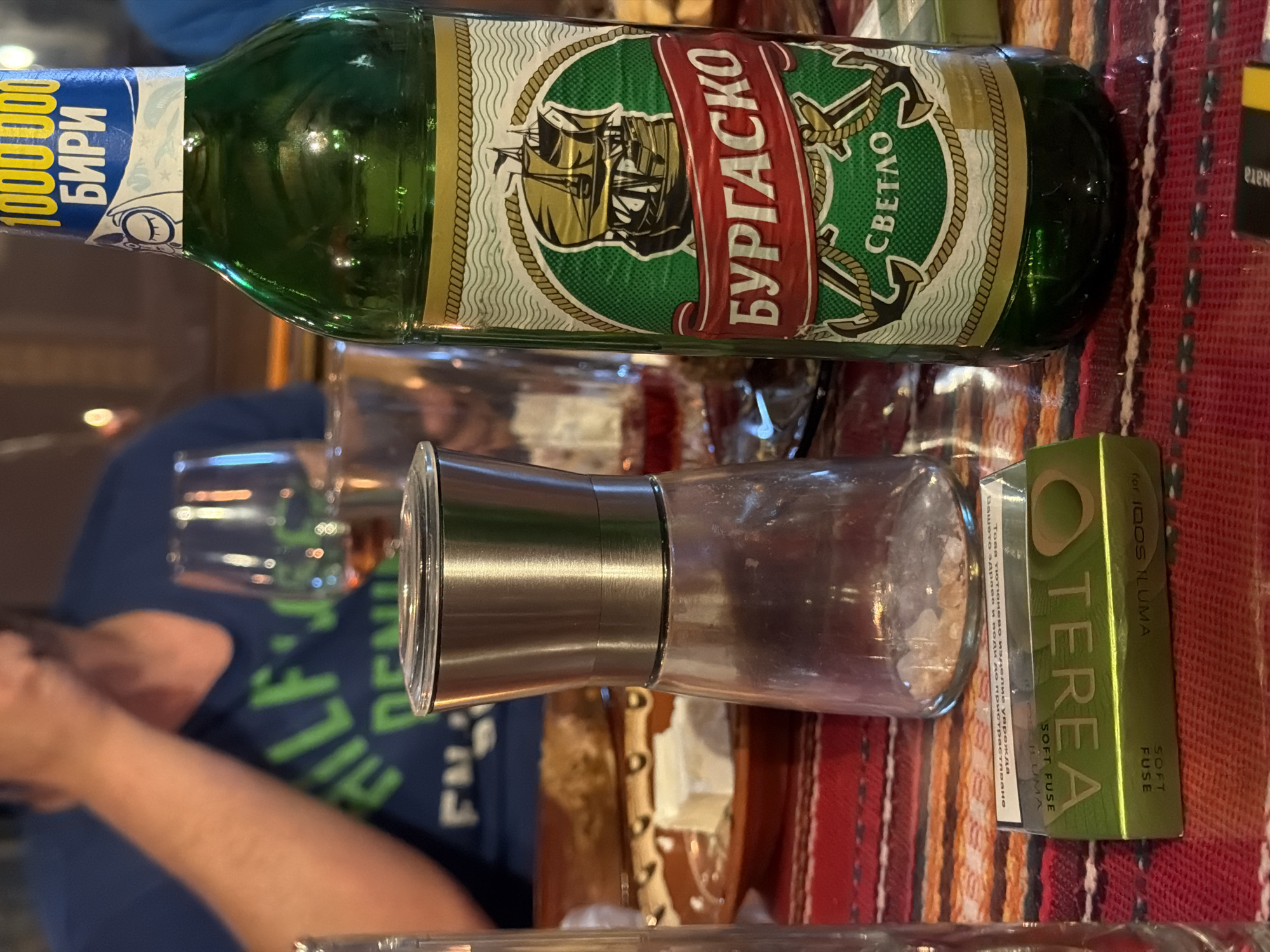 The image shows a table setting at what appears to be a restaurant or home in an unidentified location, likely Tsatsarovtsi, Bulgaria. The scene features a bottle of "Burgasko Svetlo" beer, a brand locally produced. Beside it sits a clear glass spice grinder, filled with what looks like coarse salt. Next to the spice grinder is a green package of "TEREA Soft Fuse" for IQOS ILUMA. A partially visible person is seated at the table. The tabletop is covered with a red patterned tablecloth.