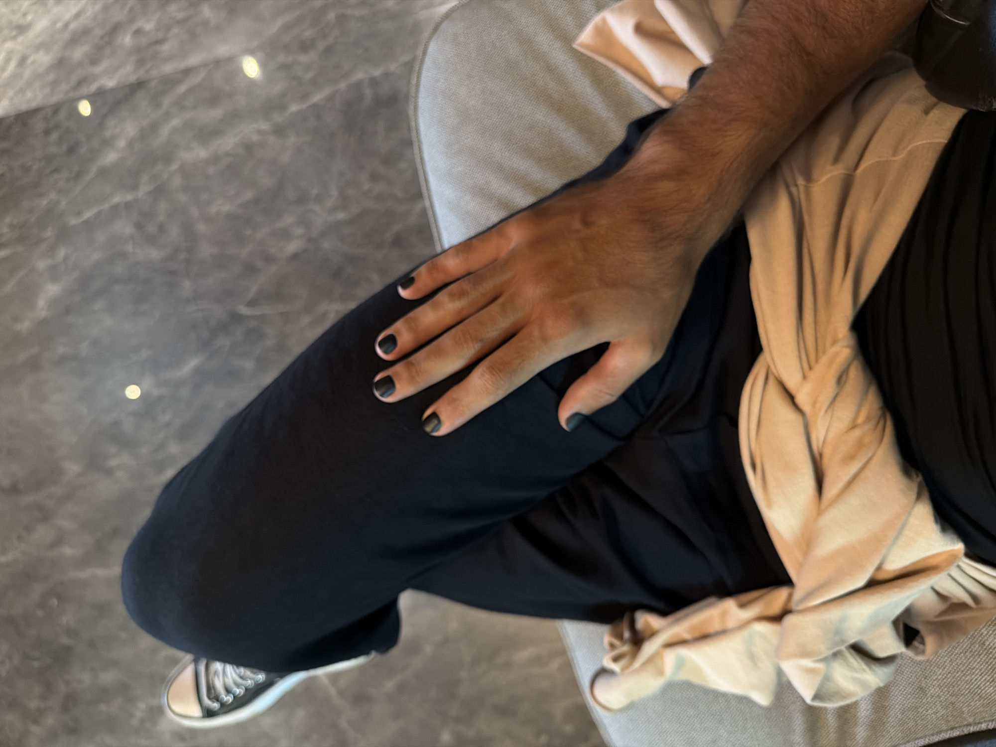 The image provides an overhead view of a person seated indoors in Kuta, Indonesia, focusing on their lap, arm, and leg. The primary subject visible is a person's right arm and hand, characterized by dark hair on the forearm and matte black nail polish on the fingernails. This hand rests on a leg clad in dark (likely black) trousers or sweatpants.
At the bottom left, a portion of a black sneaker with white laces and a white toe cap is visible. A crumpled or draped piece of light beige or tan-colored fabric, possibly a sweater or wrap, is positioned across the person's lap and around their waist area. The person appears to be seated on an upholstered surface, possibly a light grey or beige couch or chair, visible along the right edge of the frame.
The floor beneath is made of dark grey marbled tiles, featuring several small, bright, reflective spots, likely from overhead lighting. The setting appears to be a comfortable indoor environment, such as a lounge, hotel lobby, or a private residence. The activity depicted is simply sitting. No visible text is present in the image. The lighting suggests it is daytime.