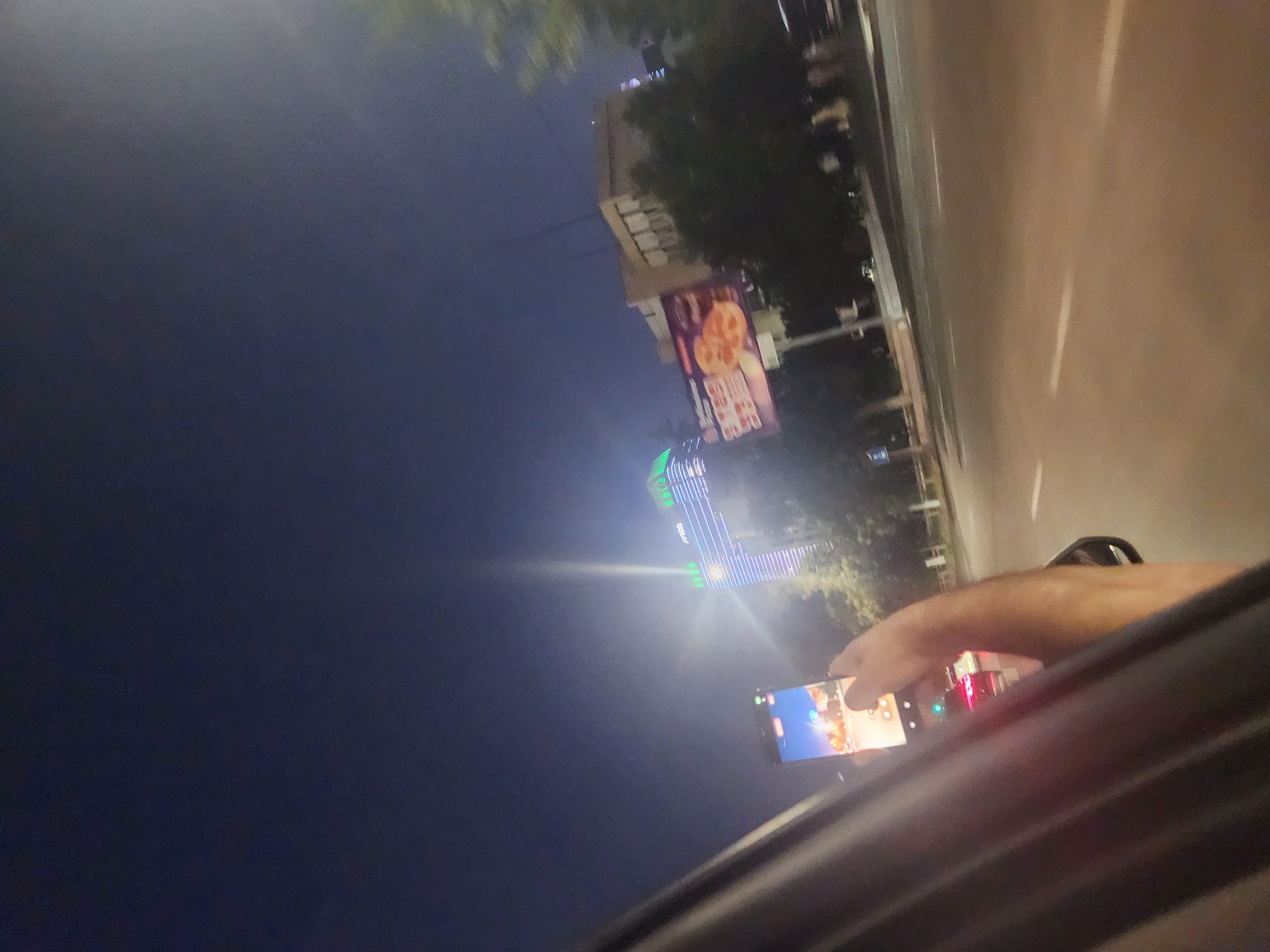 A low-angle, nighttime shot from inside a vehicle shows a hand holding a smartphone, filming a brightly lit skyscraper. The skyscraper, with its illuminated facade and green rooftop lights, is identified as the "AVIS" building, located in Tashkent, Uzbekistan. A billboard advertising pizza is visible to the right of the skyscraper. The street in the foreground is paved and has white lane markings. The sky is dark.
