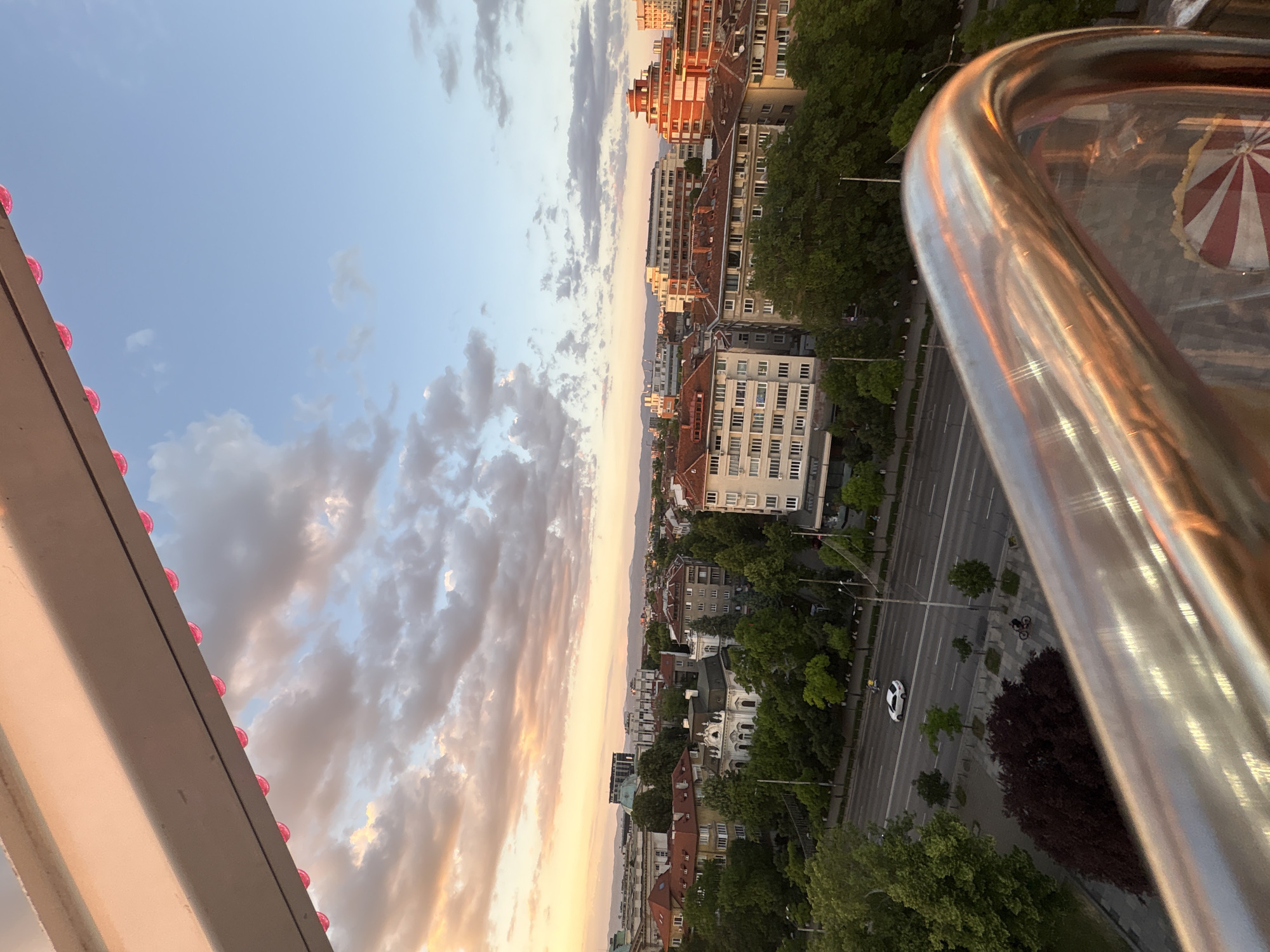 The image shows a view from a high vantage point, likely a Ferris wheel, overlooking the city of Sofia, Bulgaria. A polished metal railing is prominently featured in the foreground. The cityscape comprises buildings of varying heights and architectural styles, interspersed with green trees lining a wide boulevard with cars and bicycles. The sky has clouds in the background, indicating either sunrise or sunset. The Ferris wheel's structure, decorated with a row of pink lights, is seen on the left side of the image. The overall scene captures an urban landscape under a colorful sky.