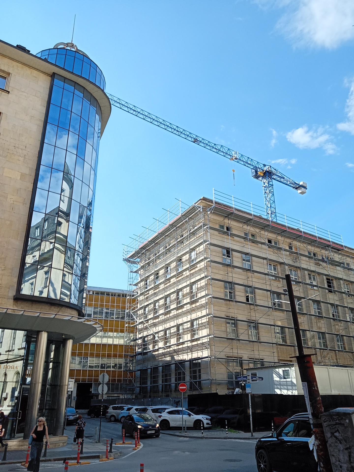 The image captures a street scene in Sofia, Bulgaria, featuring a modern building with a glass facade on the left, reflecting the sky and an older building. To the right, a multi-story building is under renovation, encased in scaffolding. A large blue construction crane extends across the frame from the background, positioned above the renovated building. Several cars are parked and driving on the street, and two pedestrians are visible walking on the sidewalk. The sky is clear blue with some scattered clouds. The scene appears to be in a commercial or mixed-use district of the city.