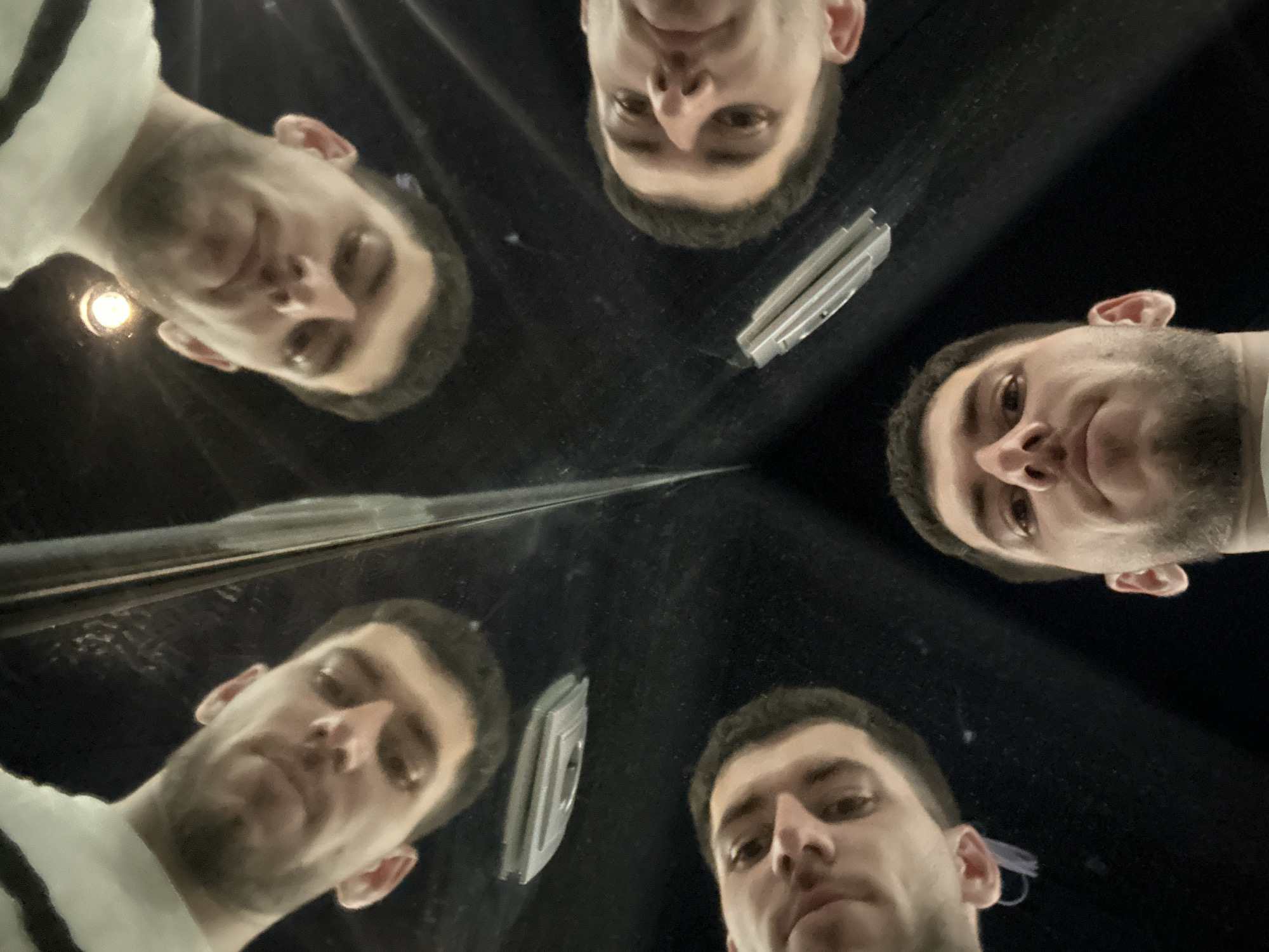 A photograph taken from a low angle, likely with a smartphone or similar device, shows a reflection of a man in a dark, possibly reflective, surface. There are multiple mirrored images of his face. An object, possibly a metallic or plastic device with a rectangular shape and a circular opening, is also reflected near the center of the frame. The environment appears dark and somewhat blurry, suggesting an indoor location with limited lighting. The specific location of the city of Sofia, Bulgaria, cannot be identified from the image alone.
