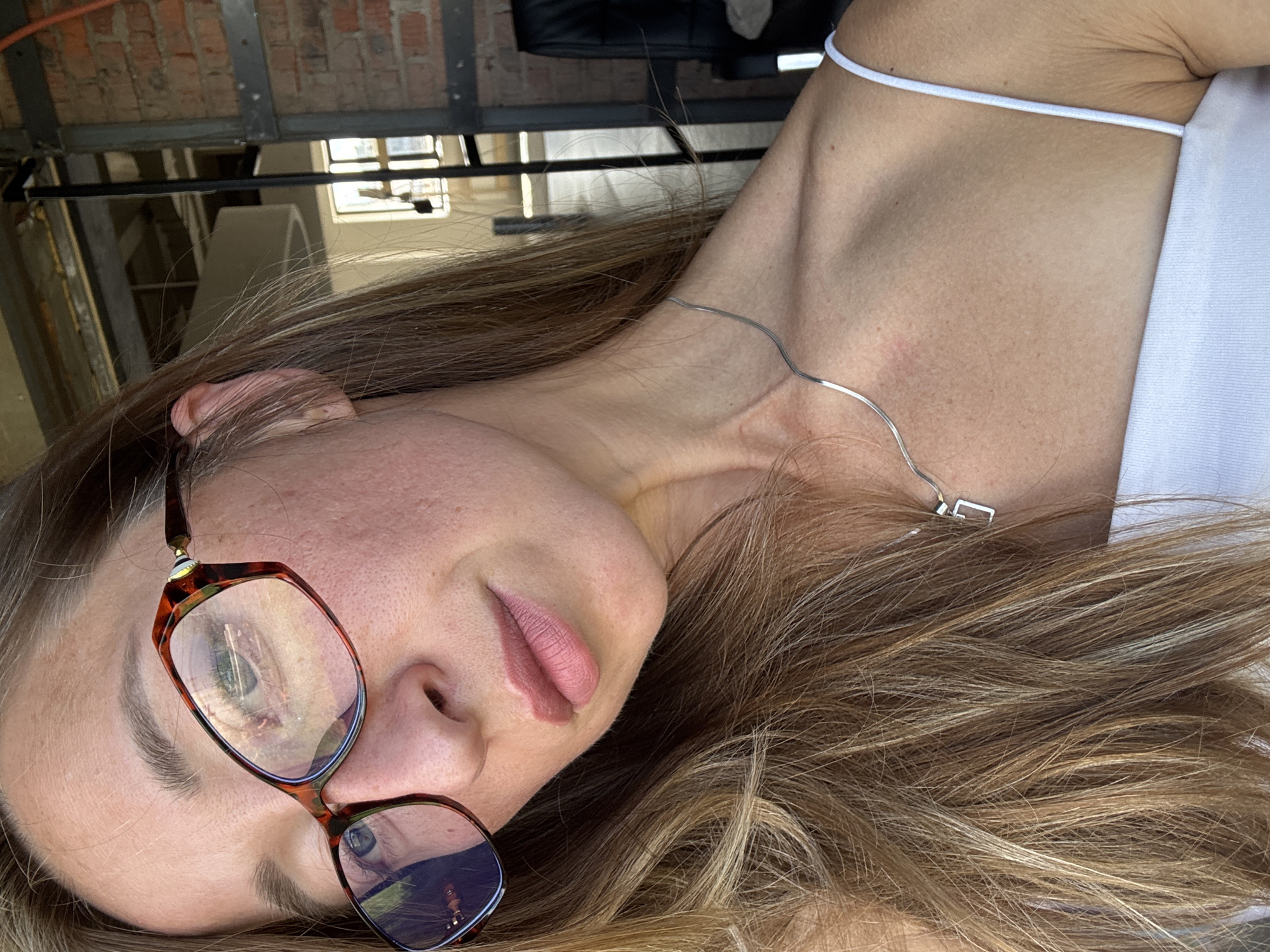 A close-up, self-portrait style photograph shows a young woman with blonde hair and fair skin, wearing glasses with tortoiseshell frames and a thin silver necklace. She is wearing a white tank top. Her face is turned slightly away from the camera, and she has pink lipstick on. The background appears to be an indoor space with exposed brick walls and industrial-style metal beams, suggesting a loft or warehouse setting, possibly in the city of Chaikovsky, Russia. There is a window with natural light coming through, and a black chair is visible in the background. The image is well-lit, and the focus is on the woman's face and upper body.