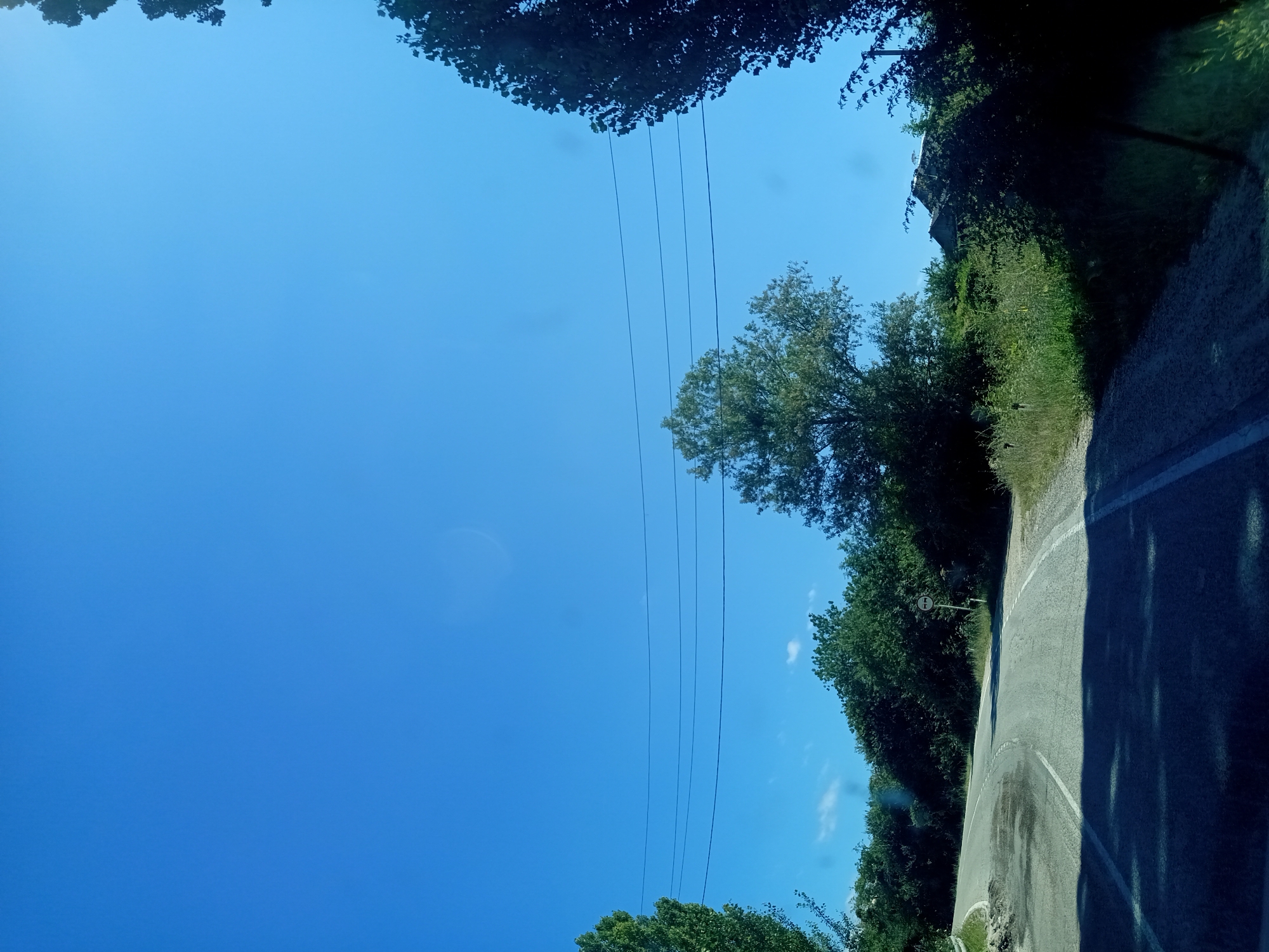 The image depicts a paved road winding through a rural landscape under a clear blue sky. Lush green trees and vegetation line the sides of the road. Several power lines are visible stretching across the upper portion of the frame. In the distance, a building is partially obscured by trees. A road sign with two circles and a horizontal line is visible on the right side of the road. The scene appears to be a sunny day in a rural area, possibly in or around Giaginskaya, Russia, given the context. The road surface shows signs of wear and tear, with some patched areas. The overall impression is one of a quiet, natural setting.