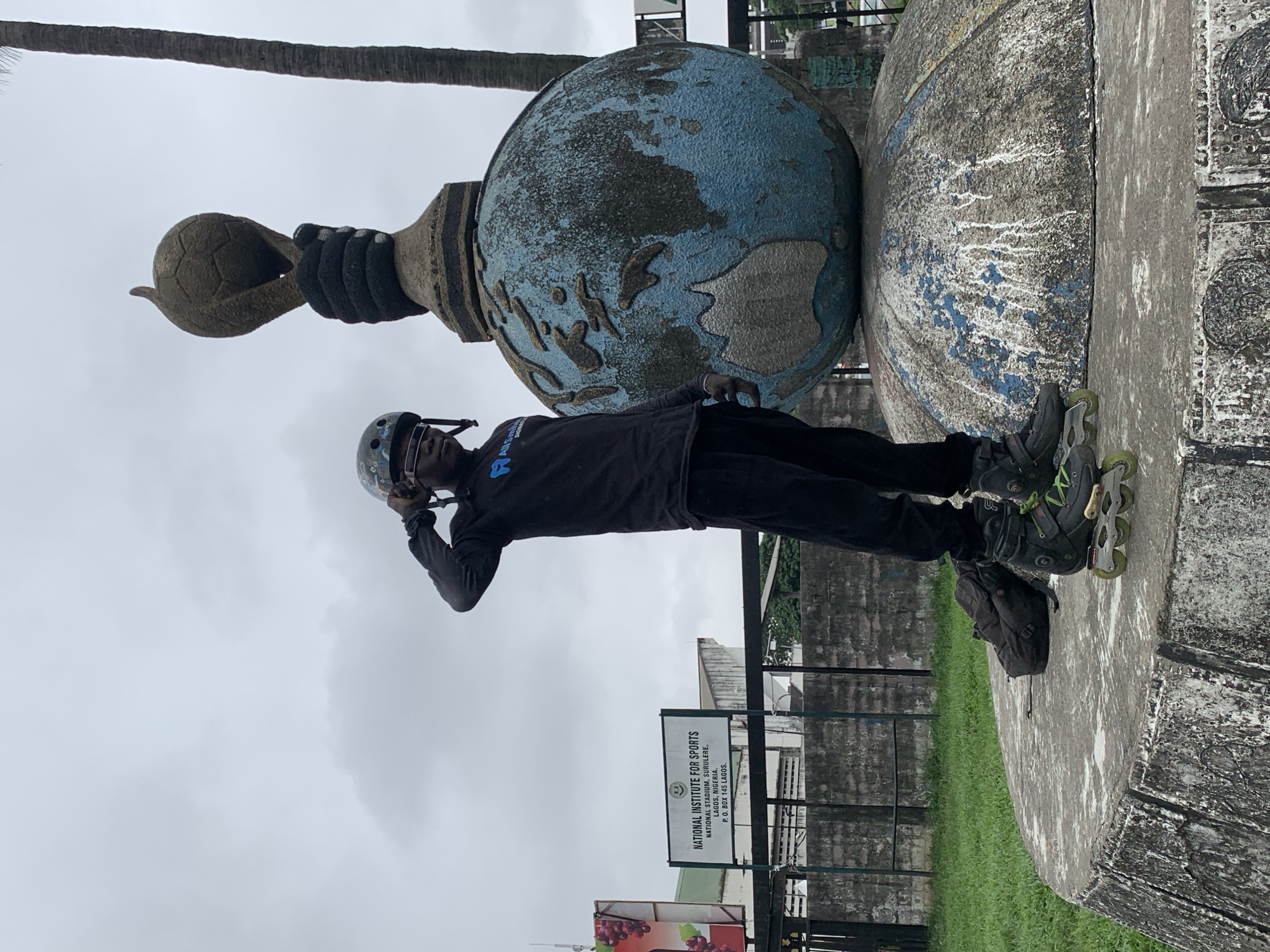 A person wearing a black long-sleeved shirt, black pants, a helmet, and rollerblades stands on a circular concrete platform. They are touching the helmet with their right hand. Behind the person is a large statue of a globe with a soccer ball held in a hand above it. The statue appears weathered and has blue and grey coloring. To the left of the person, there is a black backpack. A sign with text is visible in the background, reading "NATIONAL INSTITUTE FOR SPORTS, NATIONAL STADIUM, SURULERE, LAGOS, NIGERIA, P. O. BOX 145 LAGOS." The environment is outdoors with a cloudy sky, green grass, and a palm tree. The location is identified as the National Institute for Sports in Iganmu, Nigeria.