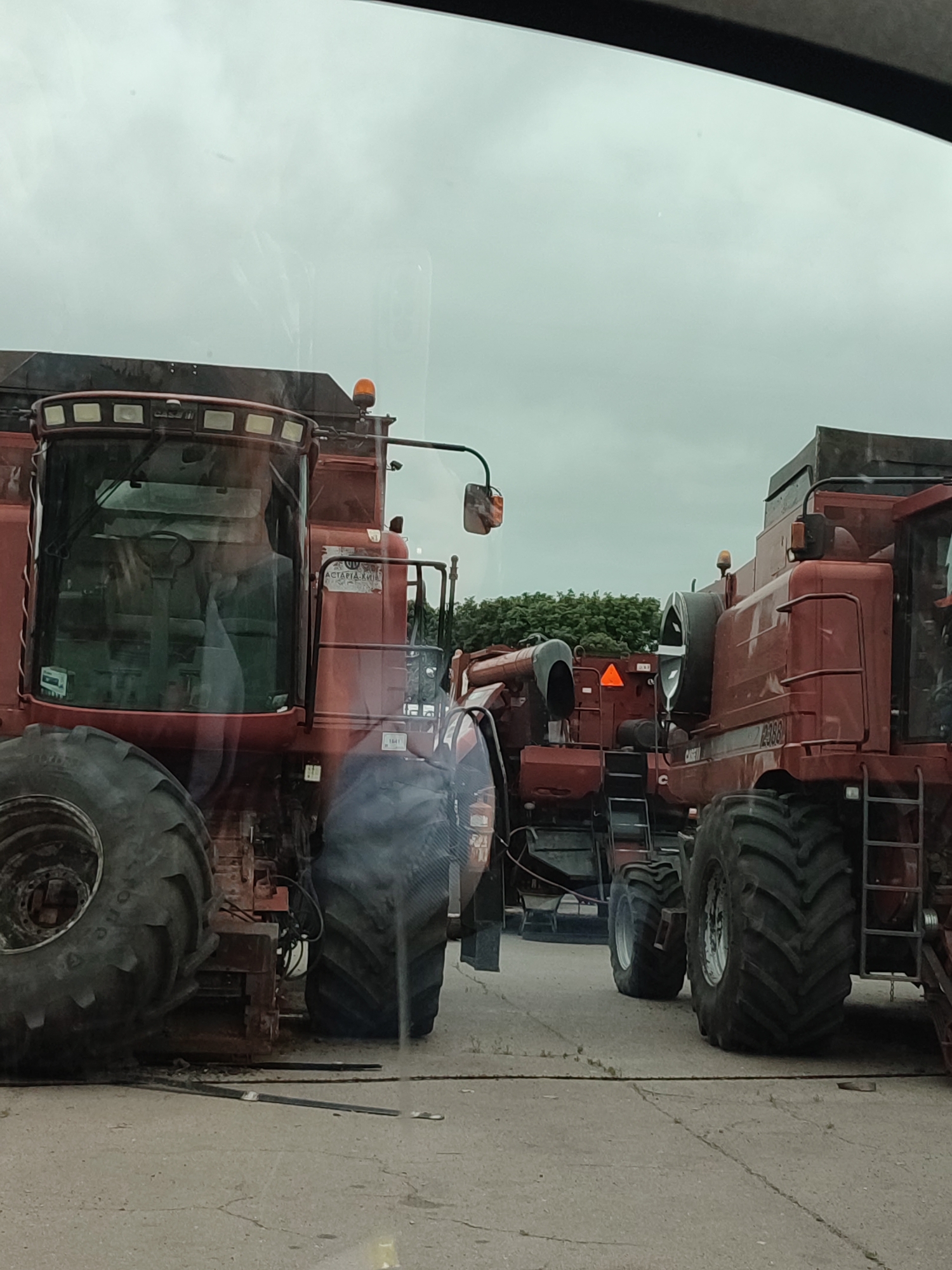 Two large red combine harvesters are parked side-by-side on a paved surface. The combine on the left is in the foreground, and its large front tire is prominent. The combine on the right is partially obscured by the first. Both vehicles appear to be agricultural machinery, with prominent tires and harvesting headers visible. The sky is overcast.
