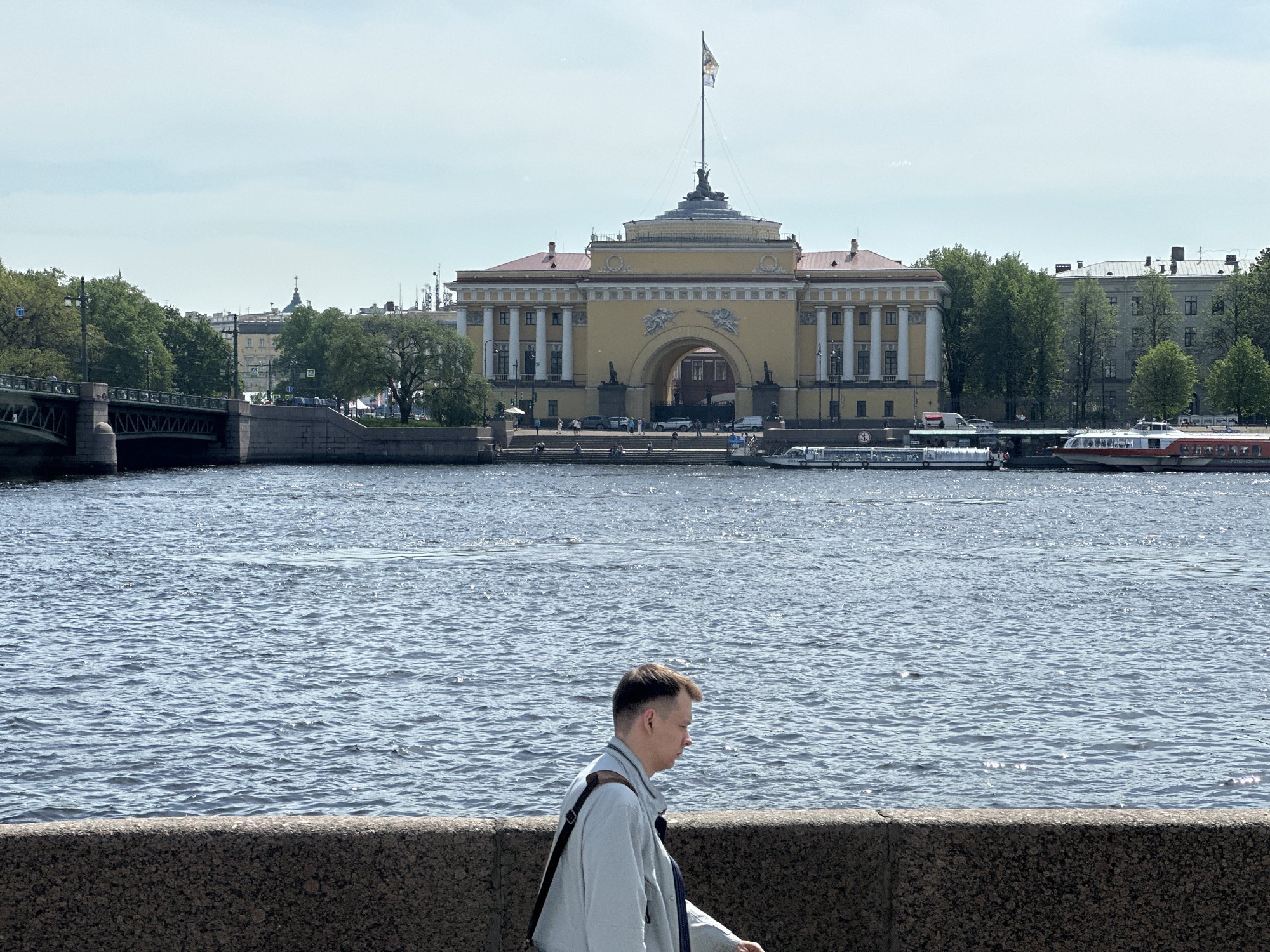 The image shows a view in Saint Petersburg, Russia, specifically the Admiralty building across the Neva River. A person is walking in the foreground along a stone embankment. The Admiralty, a large yellow building with white columns and a central archway topped with a spire and flag, dominates the background. Several boats are docked along the river in front of the building. A bridge is visible to the left of the Admiralty, and buildings and trees line the riverbank. The sky is partly cloudy.