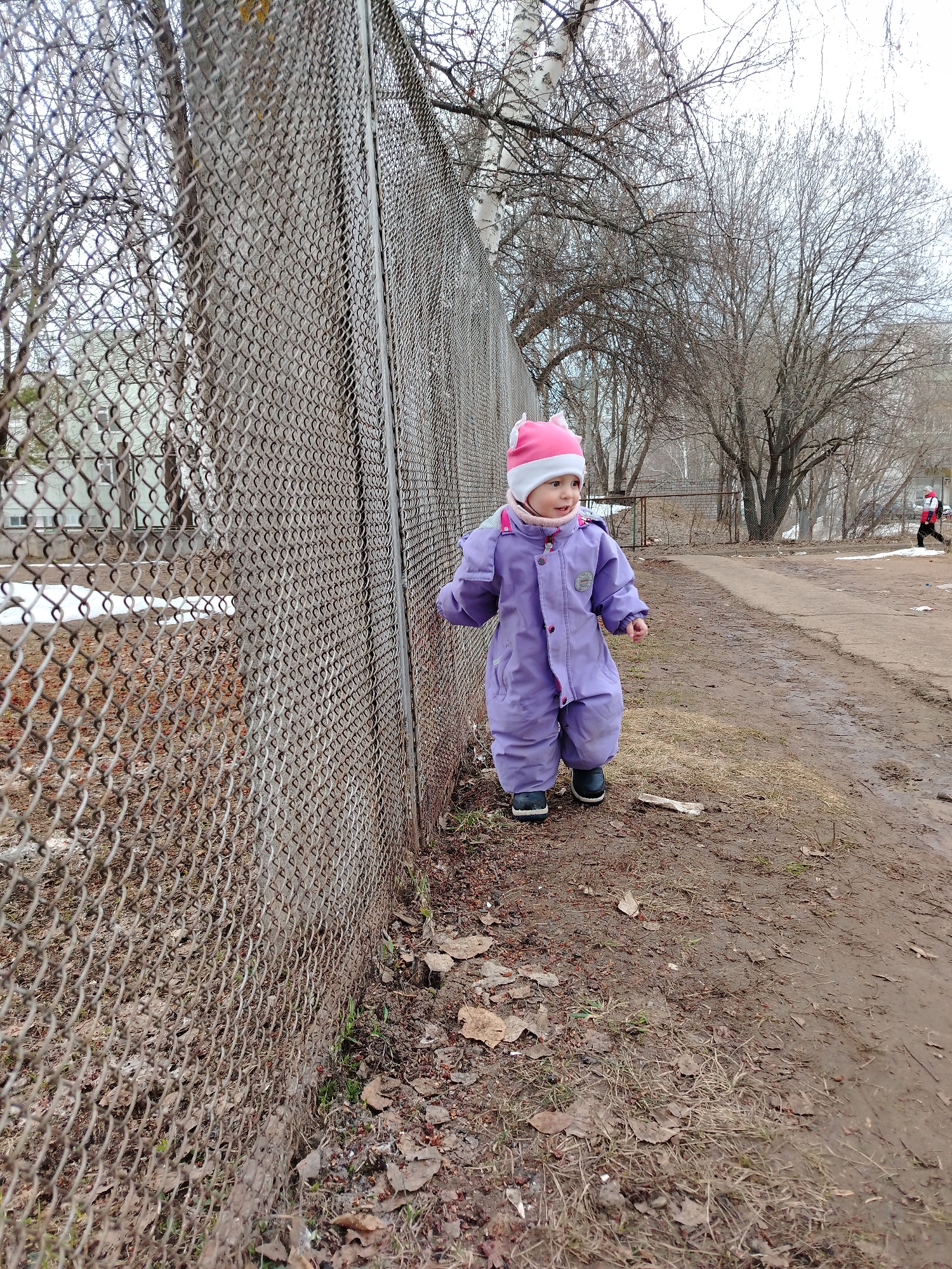 A young child wearing a purple snowsuit and a pink and white hat walks along a dirt path next to a chain-link fence. The path is lined with dry leaves and bare trees. In the background, buildings and another person can be seen. The location is likely Chaikovsky, Russia, given the request.