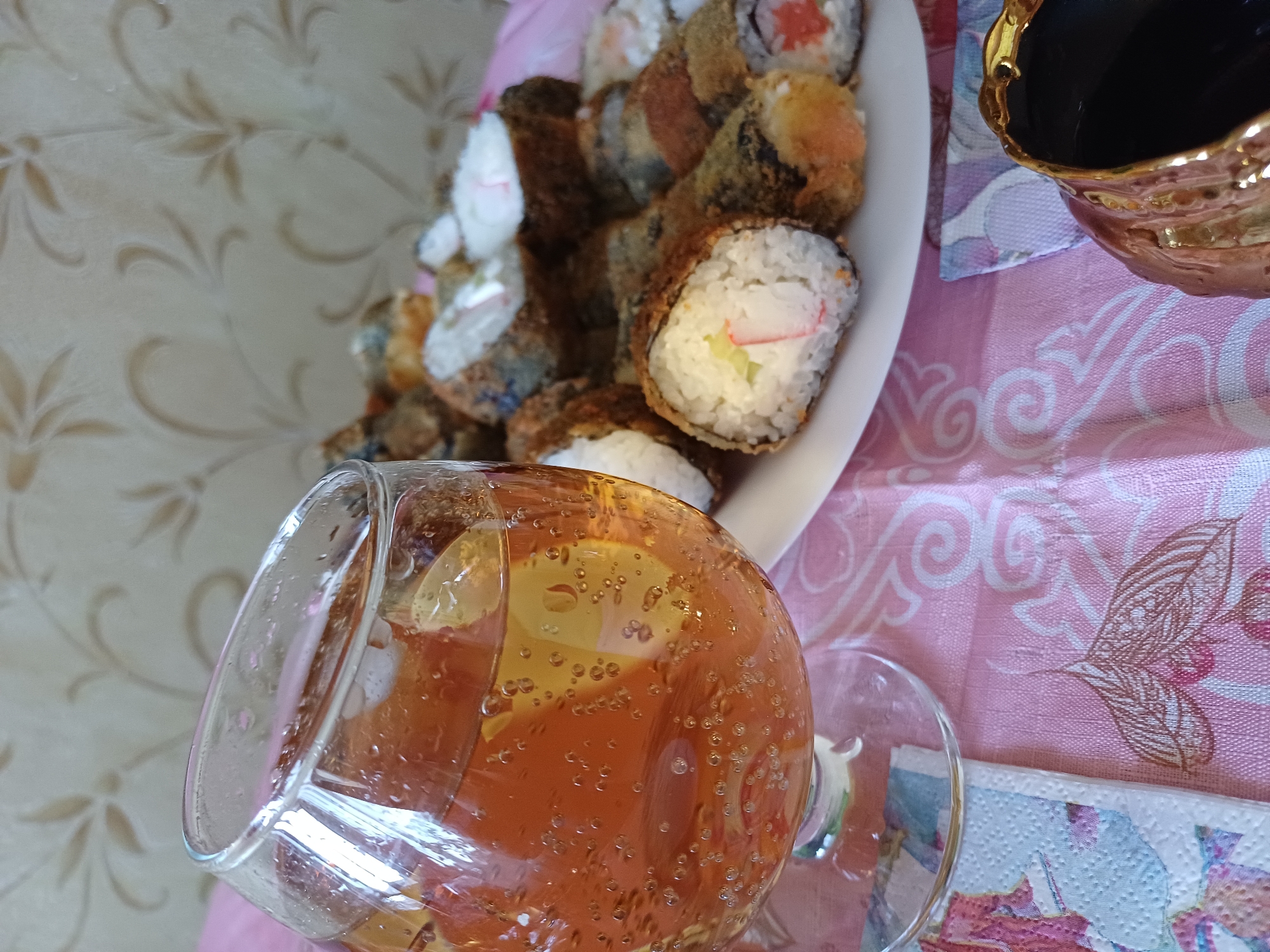 A close-up, slightly low-angle shot captures a glass of amber-colored beverage filled with small bubbles, sitting on a pink tablecloth with a floral pattern. Behind the glass, a white plate holds a collection of deep-fried sushi rolls. The rolls are varied, some showing visible fillings like cucumber and what appears to be imitation crab stick, while others are coated in a dark, crispy exterior. The background consists of a wall with a neutral-toned, floral wallpaper. A portion of a decorative, golden-rimmed dark object is visible in the bottom right corner. The lighting appears to be natural, casting soft shadows.