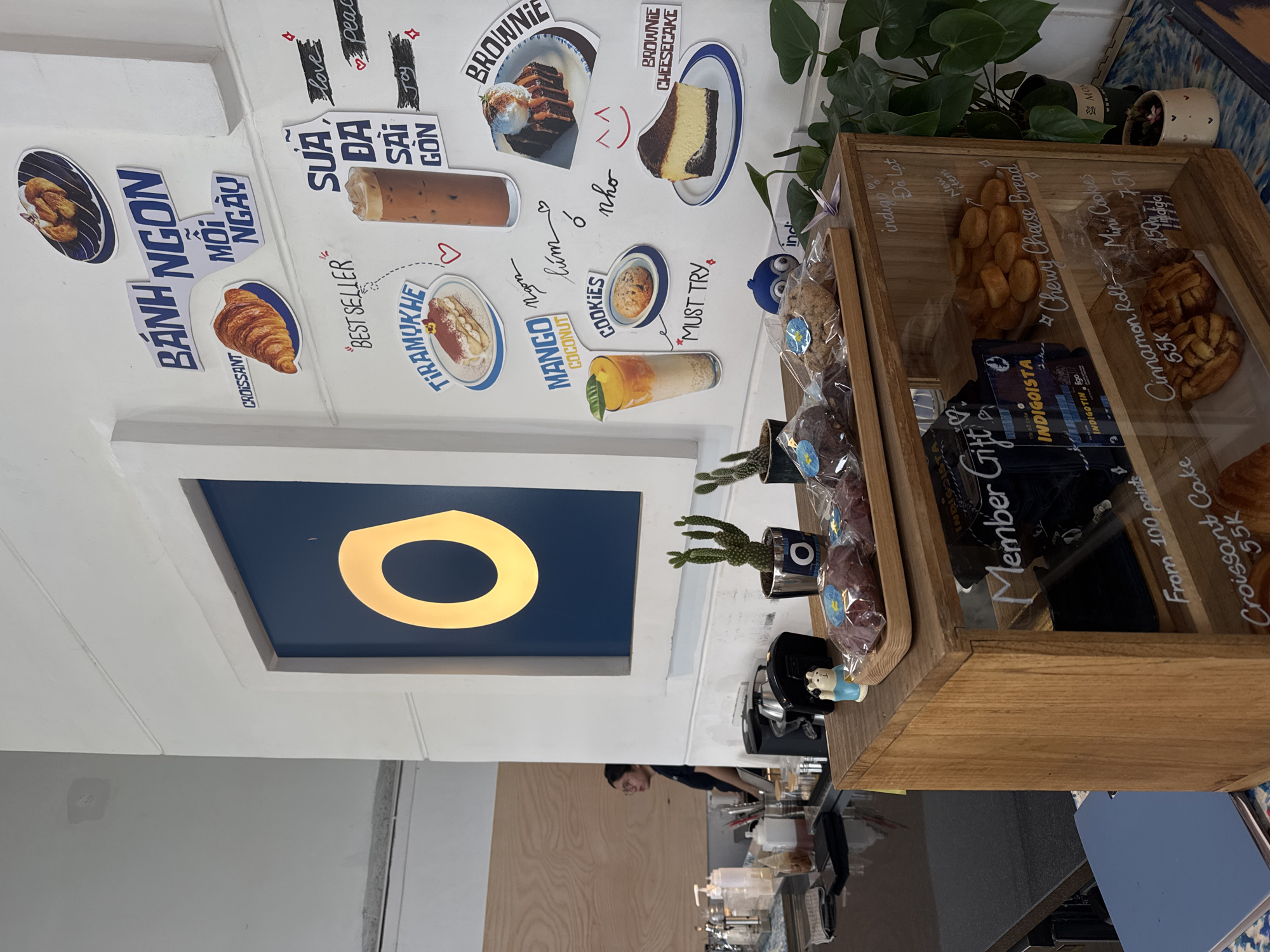 The image depicts a cafe interior, possibly located in Xuân Hương - Đà Lạt, Vietnam, given the Vietnamese text visible on the wall decorations and product labels. The scene features a white wall adorned with various posters and stickers advertising food and drinks, including a croissant, tiramisu, a mango coconut drink, brownies, and cheesecake. Text in Vietnamese, such as "BÁNH NGON MỖI NGÀY" (Delicious cakes every day) and "SỮA ĐÁ SÀI GÒN" (Saigon Iced Milk), further suggests a Vietnamese setting.
In the foreground, a wooden display cabinet is filled with baked goods. On the top shelf of the cabinet are packaged cookies and other pastries. The glass front of the cabinet displays more items, including what appears to be "Chewy Cheese Bread," "Cinnamon Roll," and "Mini Cookies," with prices in Vietnamese Dong (K).
To the left, a counter area is visible with various coffee-making equipment, dispensers, and a person wearing glasses and a dark shirt standing behind it, seemingly an employee. Small potted plants, including a cactus, are placed around the display cabinet, adding a touch of greenery to the scene. The overall atmosphere appears to be that of a modern, well-lit cafe.