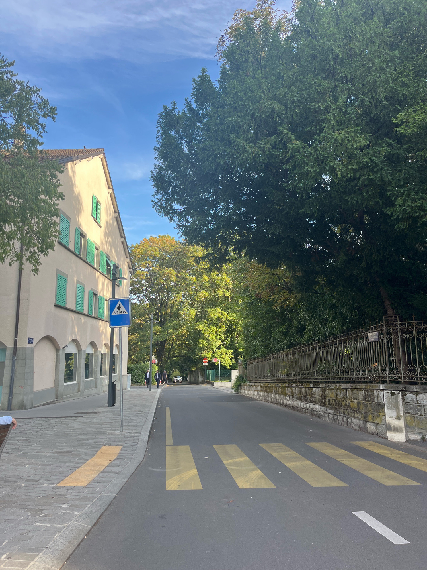 The image depicts a street scene in Geneva, Switzerland, likely in the autumn given the foliage colors. On the left is a light-colored building with several windows and green shutters. The building appears to be multi-story. In front of the building is a sidewalk paved with gray stones, with a yellow tactile paving strip near the curb. A blue pedestrian crossing sign with a white silhouette of two people walking is visible on a pole. To the right of the street is a decorative metal fence with stone foundations, bordered by lush green trees. The street itself has a pedestrian crossing marked with yellow stripes. Several people are walking on the sidewalk in the distance, and a white car is visible on the road. The sky is blue with some wispy clouds.