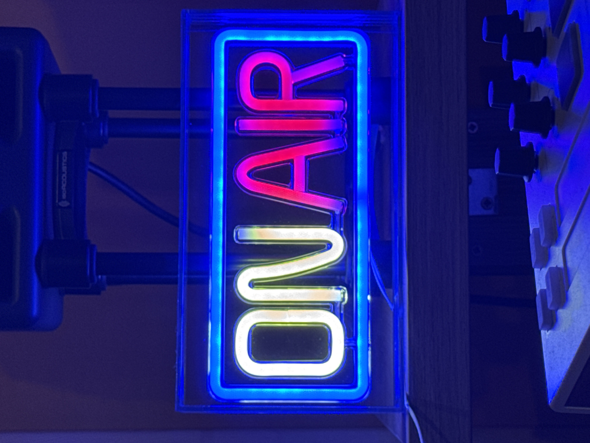 A vertically oriented neon sign prominently displays the words "ON AIR" in a dark, interior setting, likely a broadcast or recording studio in Plovdiv, Bulgaria. The sign itself features a bright blue neon border, with the word "AIR" illuminated in vivid pink-red neon and the word "ON" glowing in white-yellow neon. It is enclosed within a clear rectangular casing, suggesting a modern or well-maintained setup.
To the left of the sign, partially visible, is a piece of dark audio equipment, possibly a speaker or an isolation stand, with the brand name "ISOACOUSTICS" printed vertically in white. Black cylindrical components, likely parts of a stand or mount, connect this equipment.
On the right side of the sign, a section of what appears to be a mixing board or control panel is visible. It features several black rotary knobs arranged vertically on a lighter-colored surface, along with some lighter square buttons or sliders.
The overall lighting of the scene is dim, with the neon sign providing the primary light source, casting a blue hue onto the surroundings. The presence of professional audio equipment and the "ON AIR" sign strongly indicate an active recording session, live broadcast, or podcast in progress. No people are visible, emphasizing the equipment and the functional nature of the space.