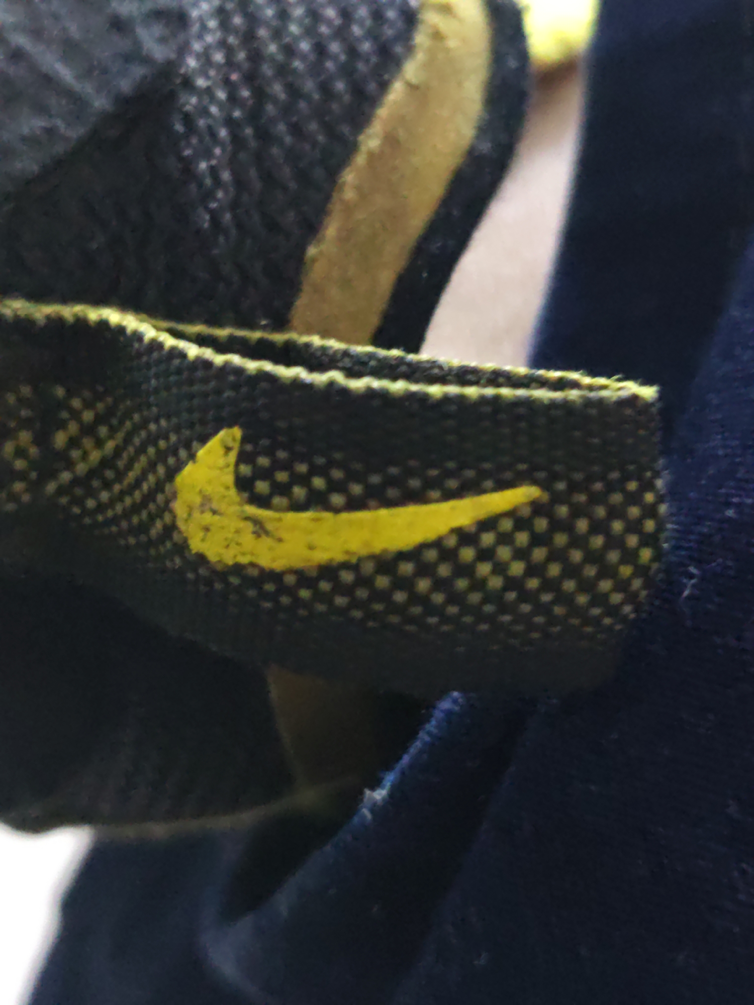 A close-up, low-angle shot shows a yellow Nike swoosh logo on a black fabric strap. The strap has a woven pattern of small white dots against the black background, creating a checkered effect. A yellow stripe is visible above the strap, and the fabric appears to be part of an athletic shoe. The background is out of focus, with dark blue fabric to the right and a lighter, blurry area to the left. The image is grainy and lacks specific environmental context of Piloncito, Venezuela.